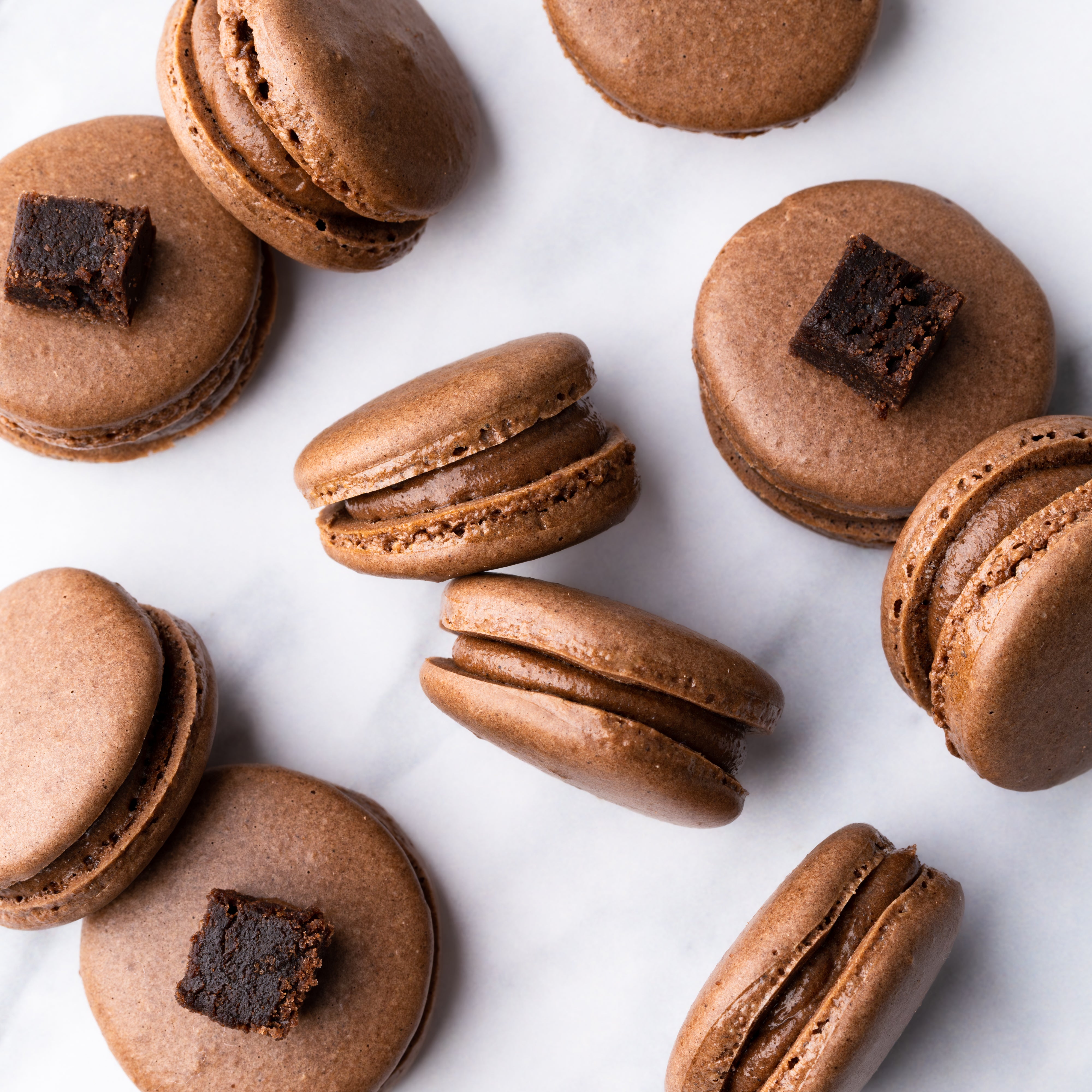 Chocolate macarons with brownie pieces on top, arranged on a white marble surface.