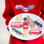 Child holding a plate of London-themed cookies, including designs of a bus, guard, Big Ben, and crown, from Craft & Crumb's baking kits for kids.