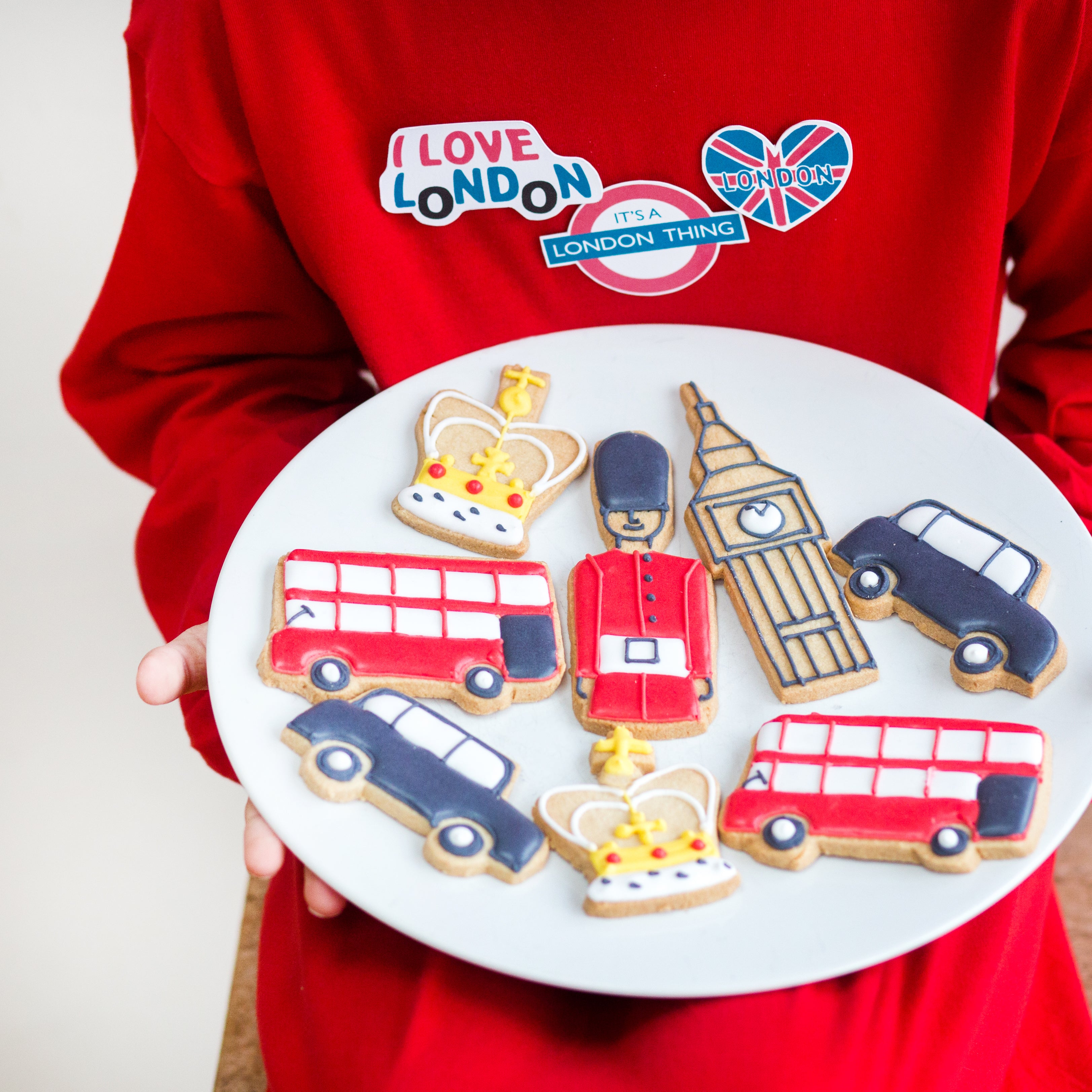 Child holding a plate of London-themed cookies, including designs of a bus, guard, Big Ben, and crown, from Craft & Crumb's baking kits for kids.