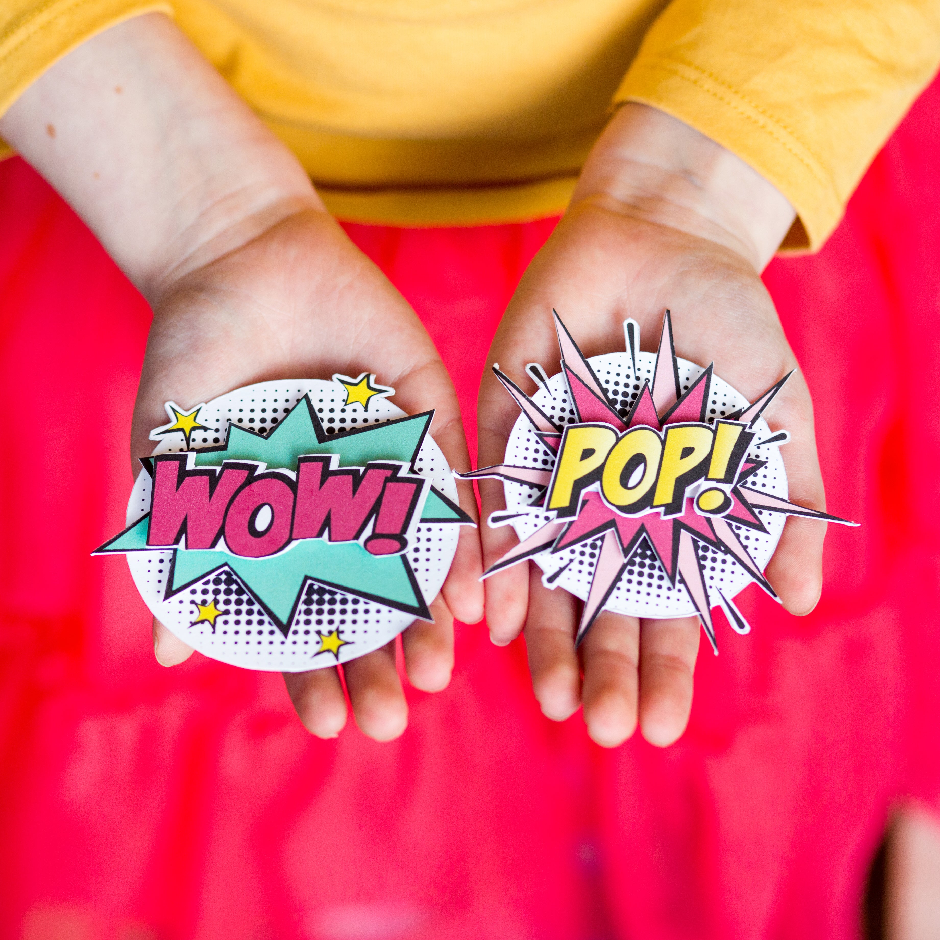 Hands holding comic-style cutouts with "WOW!" and "POP!" text, against a bright red and yellow background.