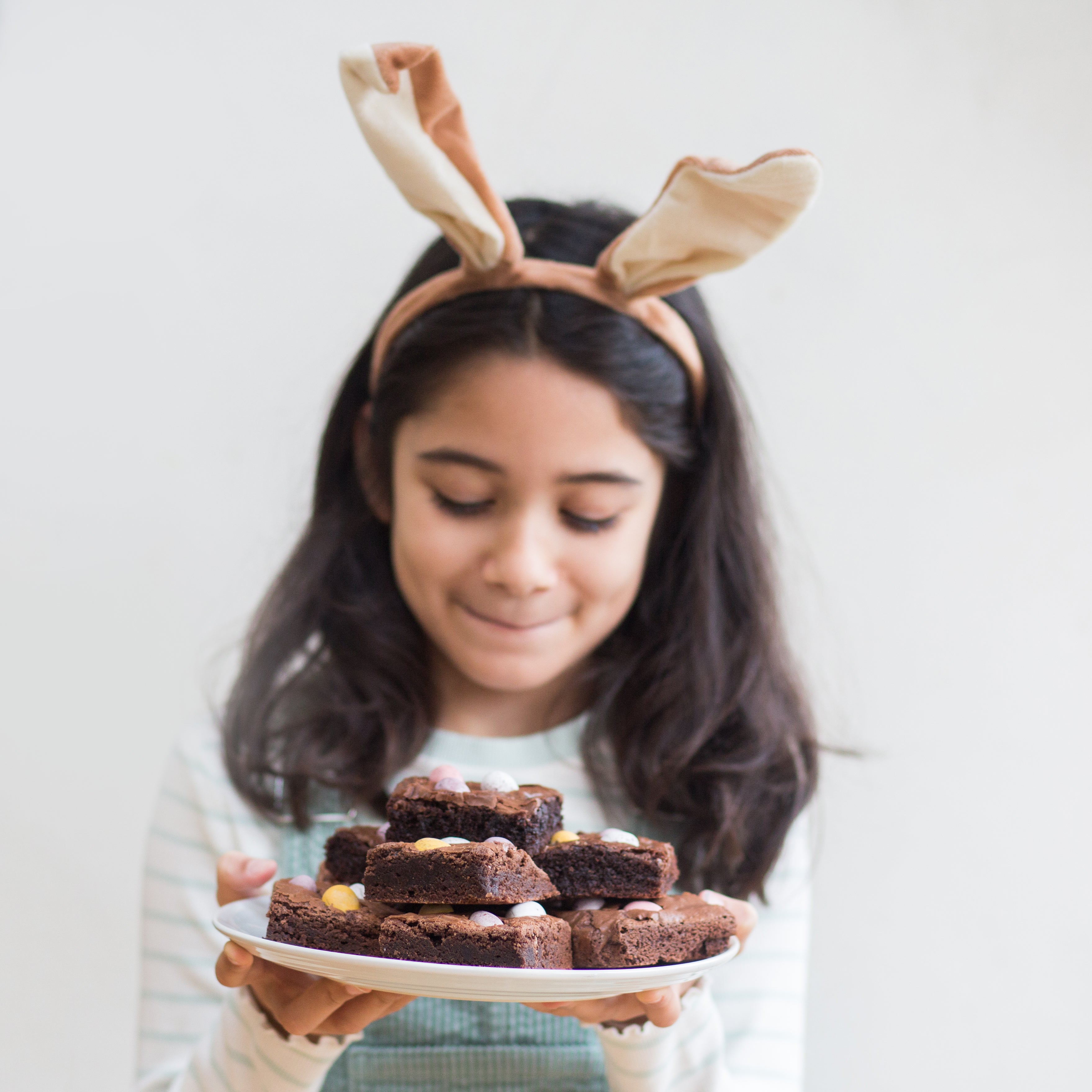 Girl with bunny ears holding a plate of brownies, smiling softly.
