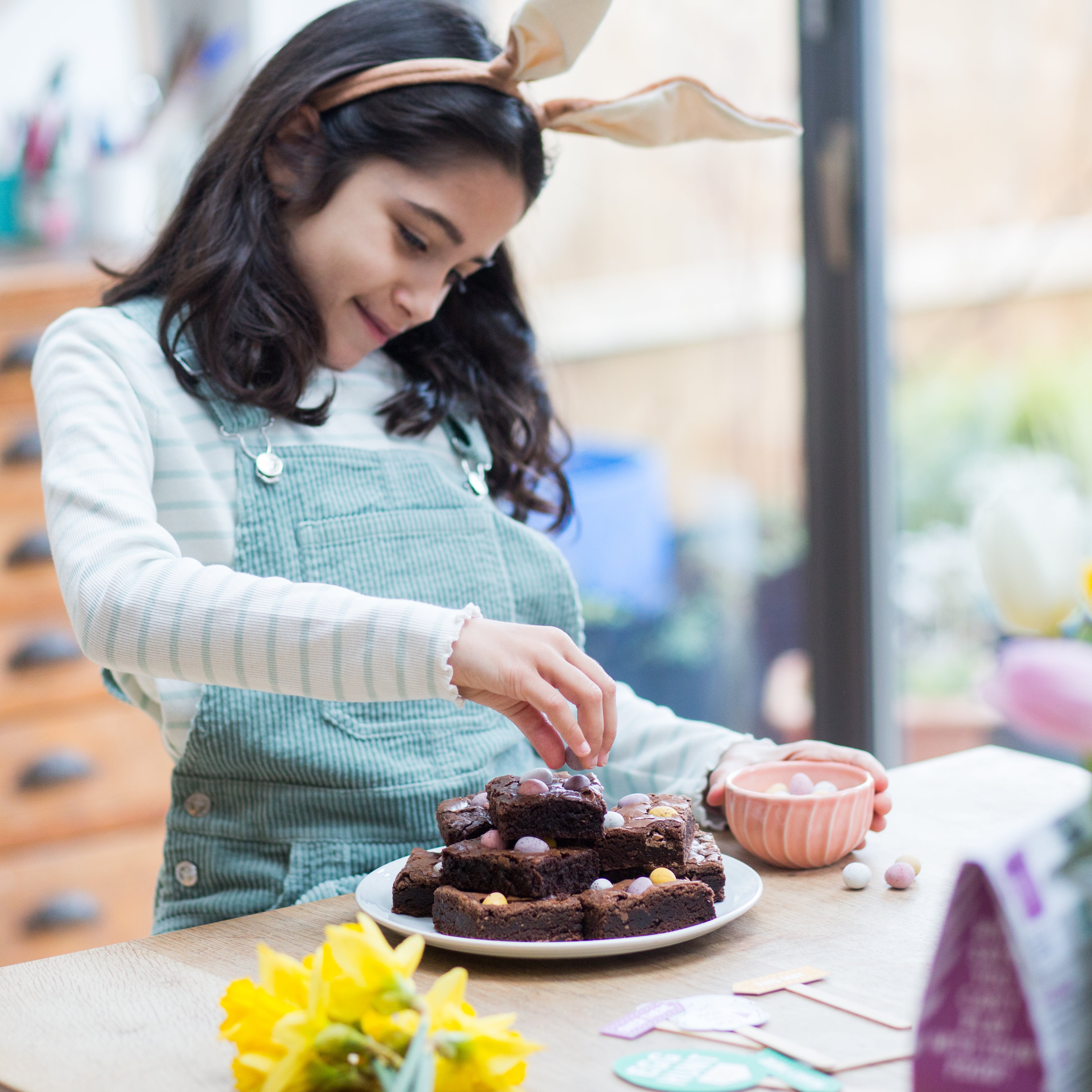 Young girl in bunny ears decorates chocolate brownies with mini eggs at a table, surrounded by spring flowers.