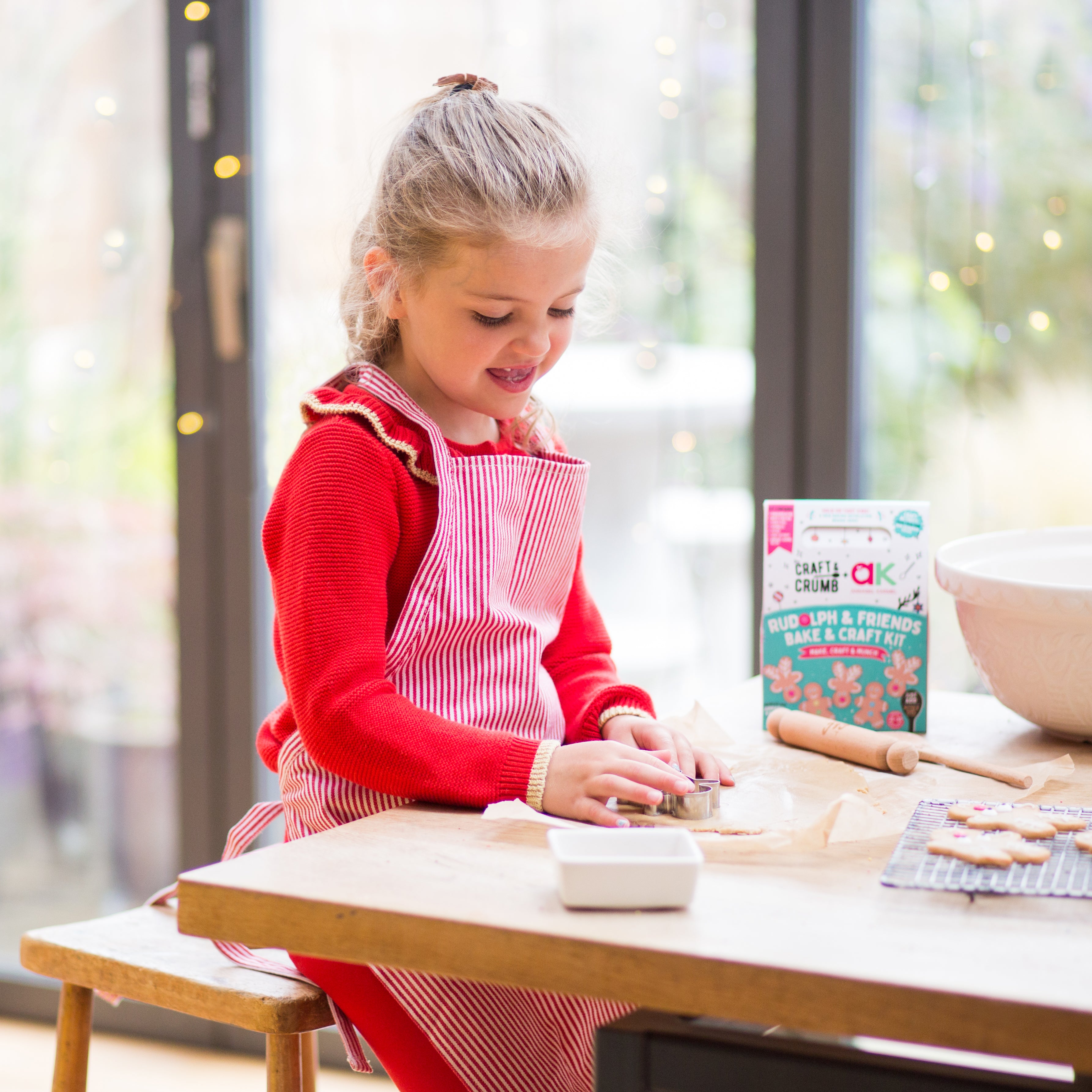 Child in red apron making cookies at a table, with a baking kit box and mixing bowl nearby, in a bright kitchen.