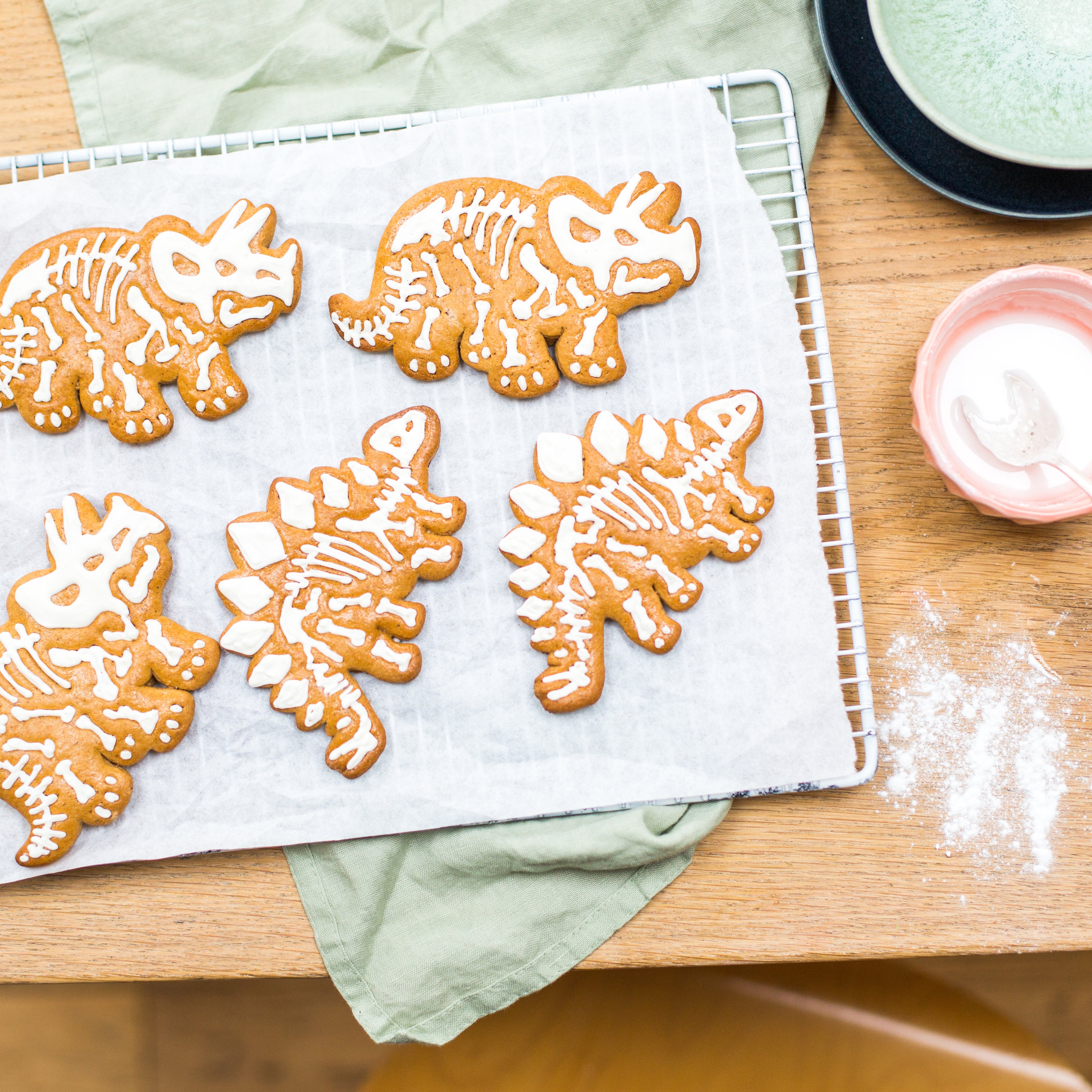 Dinosaur-shaped cookies with white icing on a cooling rack, next to a bowl of icing and a green cloth.