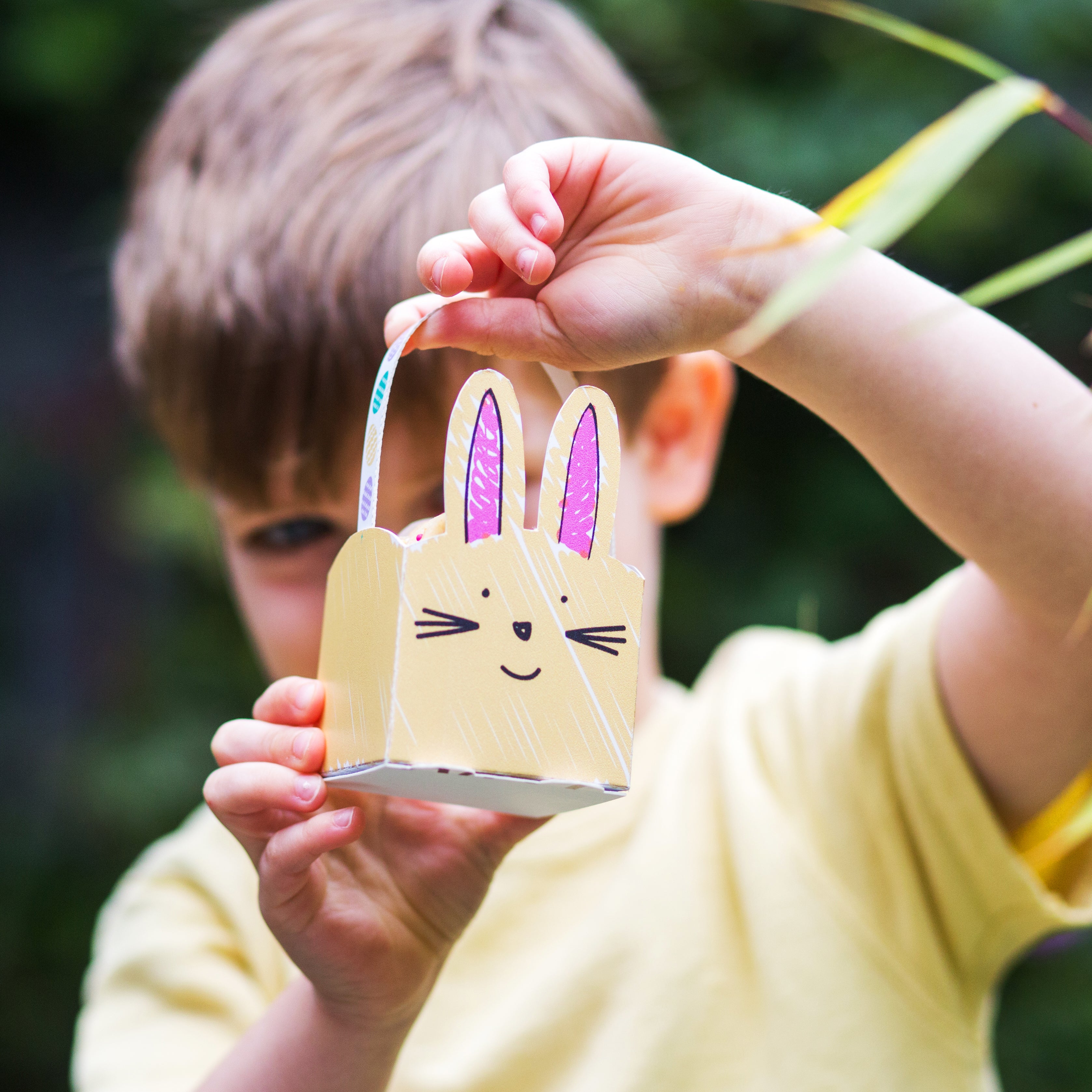 Child holding a small, bunny-themed paper basket with pink ears and whiskers, outdoors.