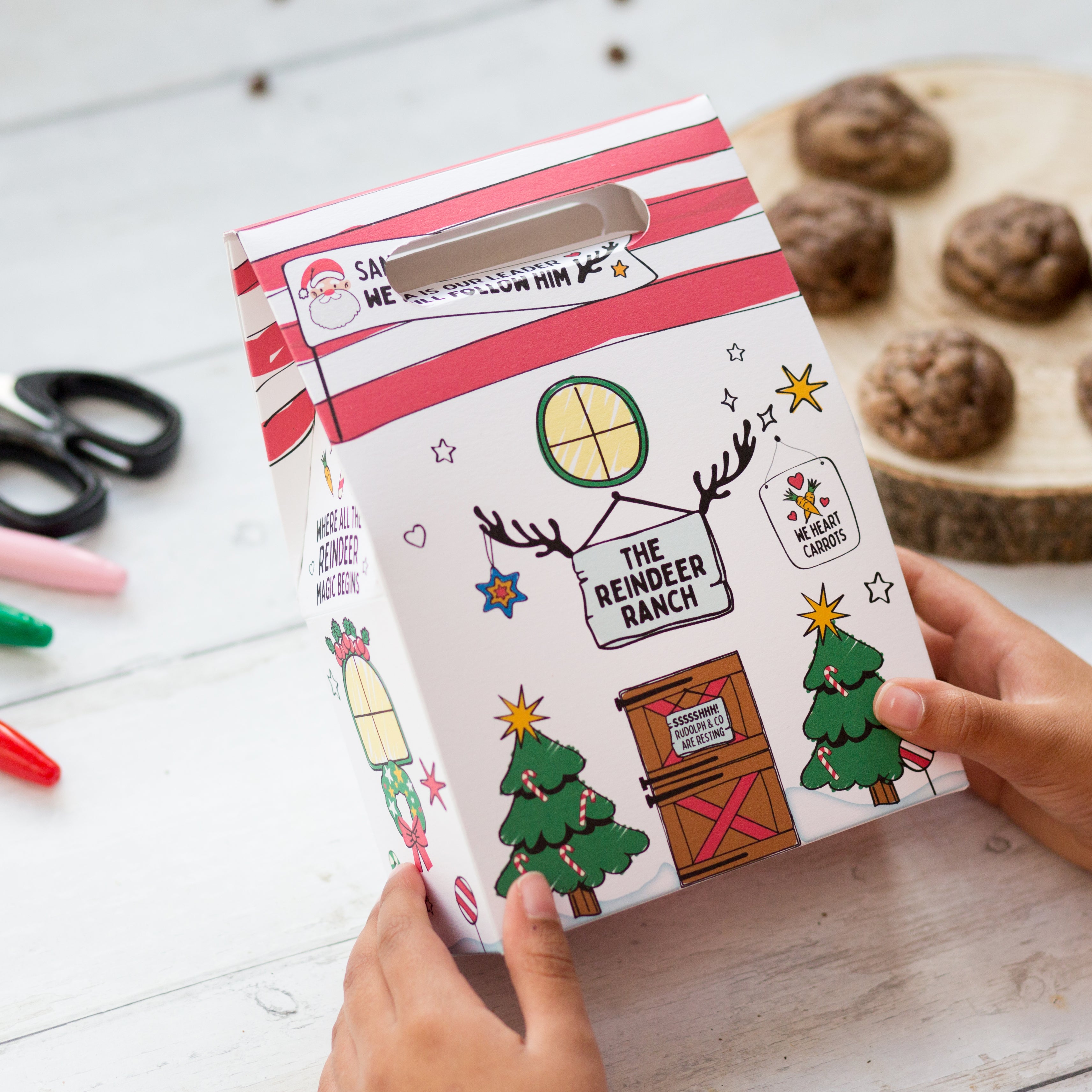 Child decorating a festive box with reindeer and Christmas tree designs, cookies and markers on a white table.