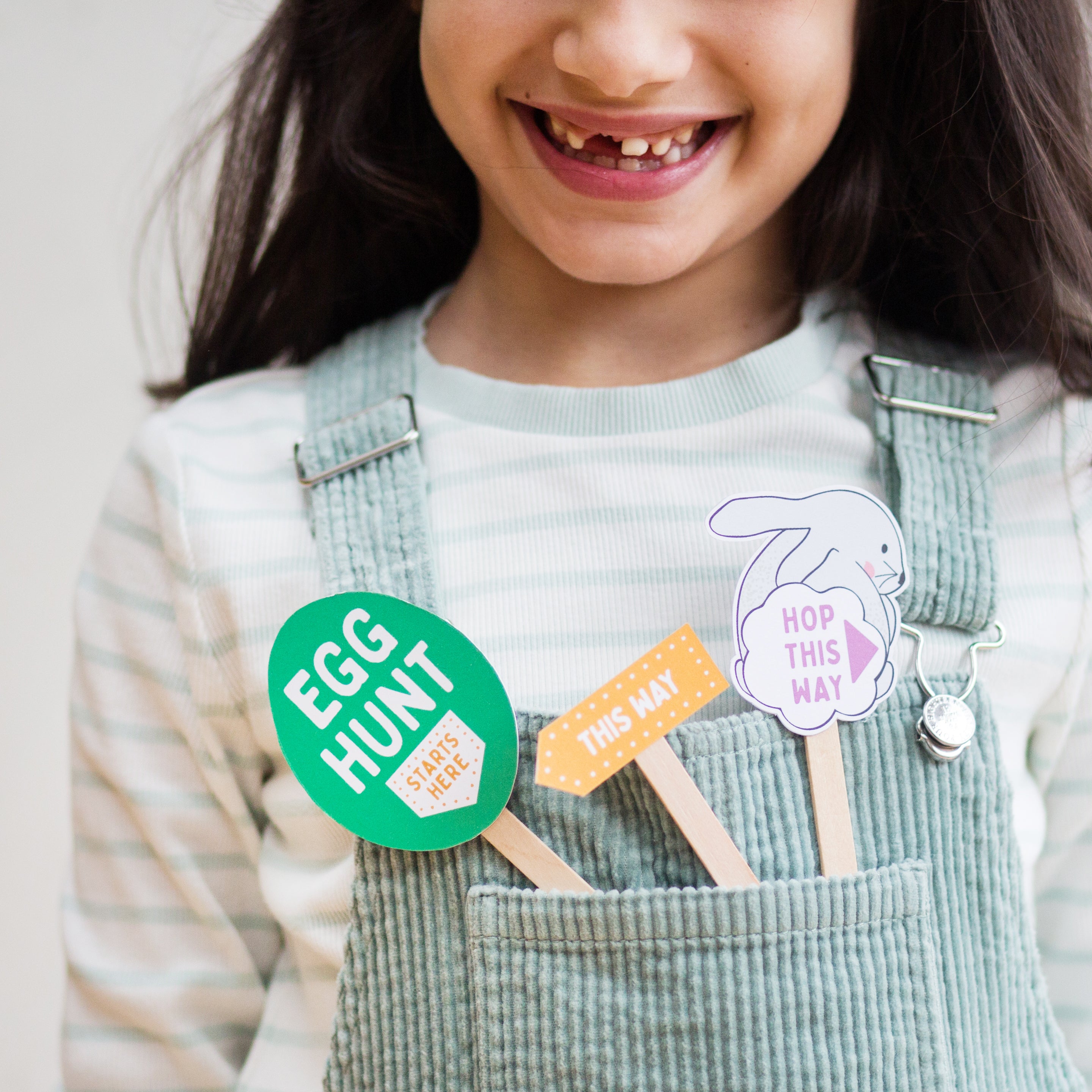 Smiling child in striped shirt and overalls holding Easter-themed signs with phrases like "Egg Hunt" and "Hop This Way."
