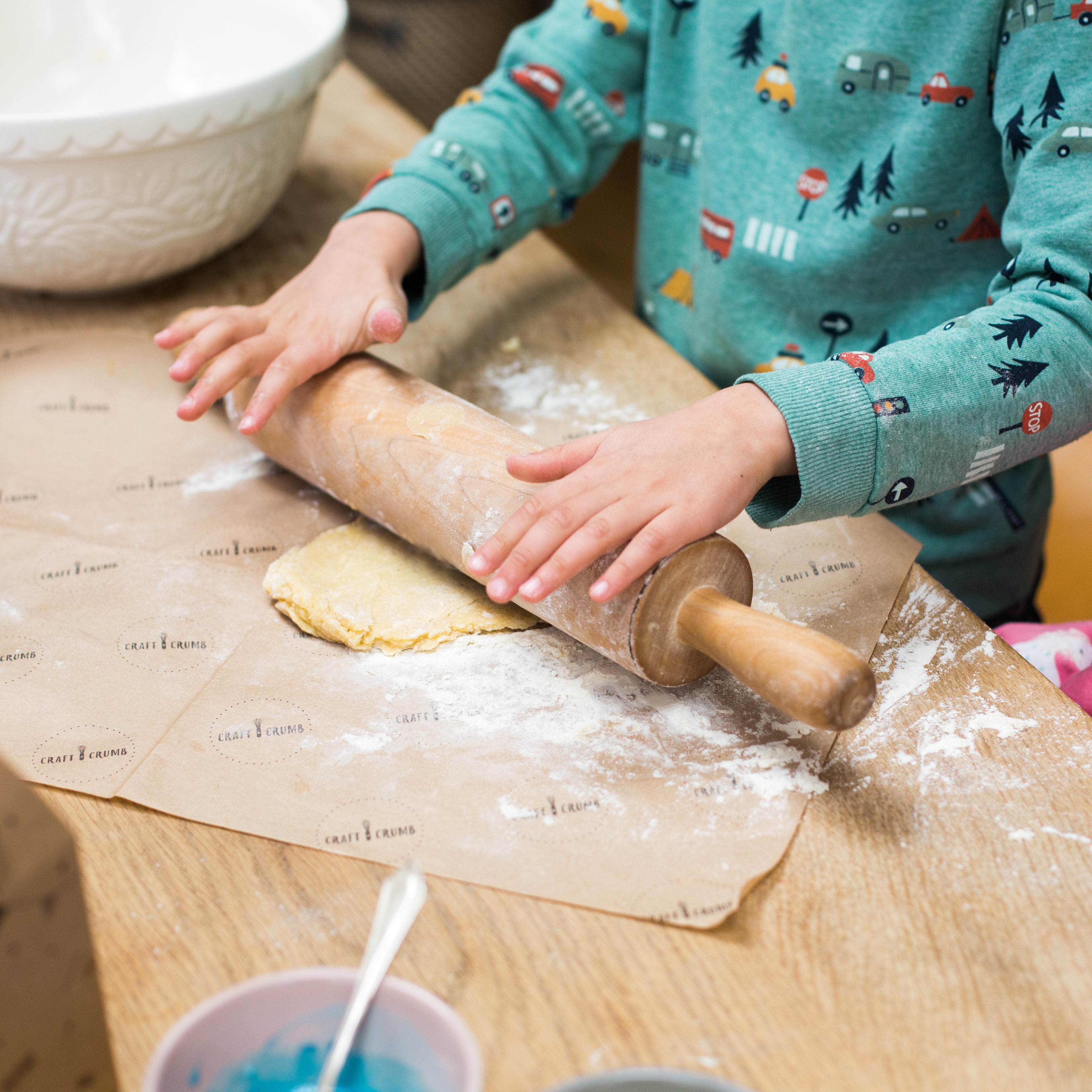 Child rolling dough with a wooden rolling pin on a floured surface, wearing a colorful sweater.