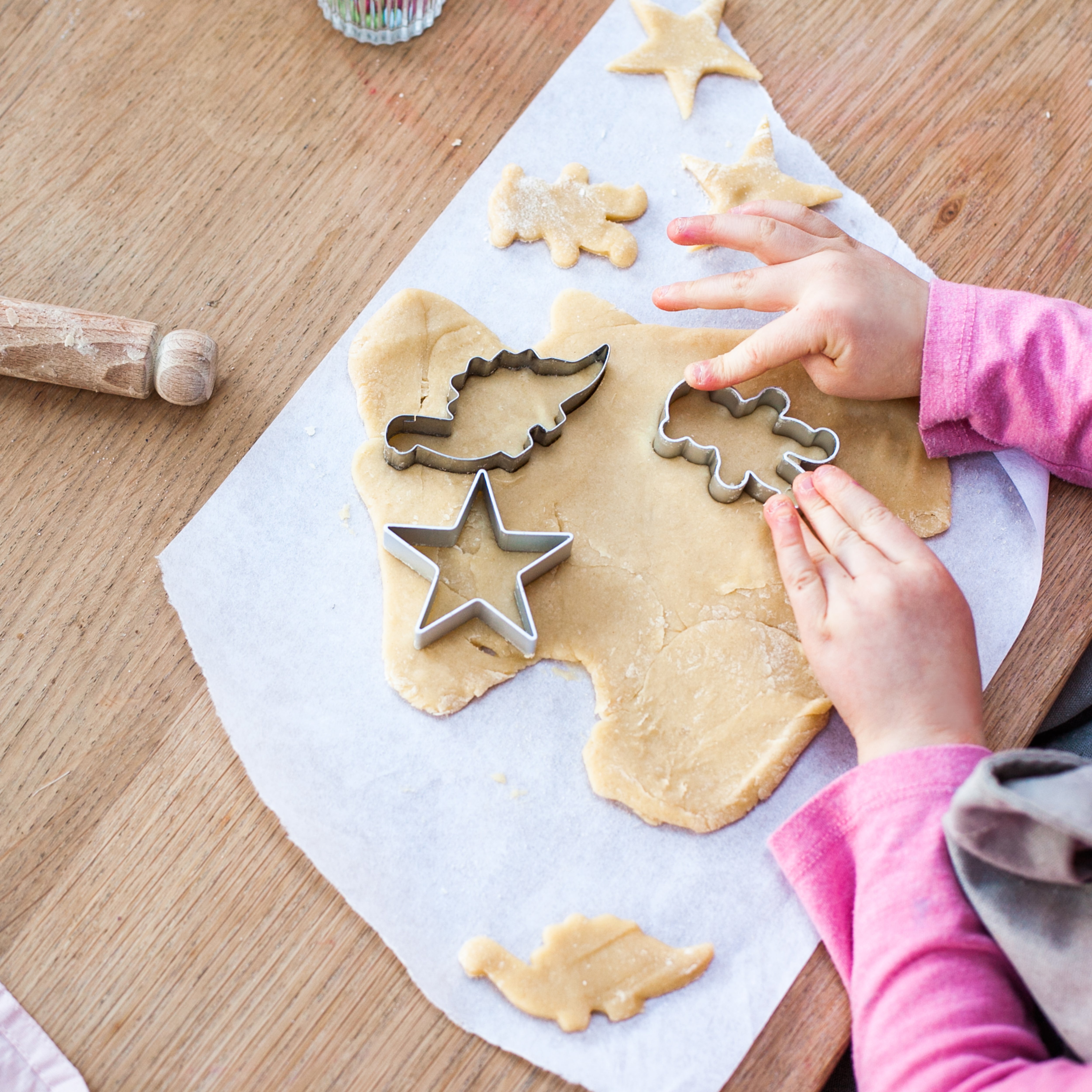 Child using cookie cutters on dough, with star and leaf shapes, on a wooden table with parchment paper.