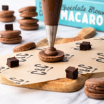 Piping chocolate filling onto a macaron shell with brownies and macarons in the background.