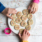 Hands reaching for sprinkle-covered cookies on a white plate, with scattered sprinkles and a pink bowl nearby.