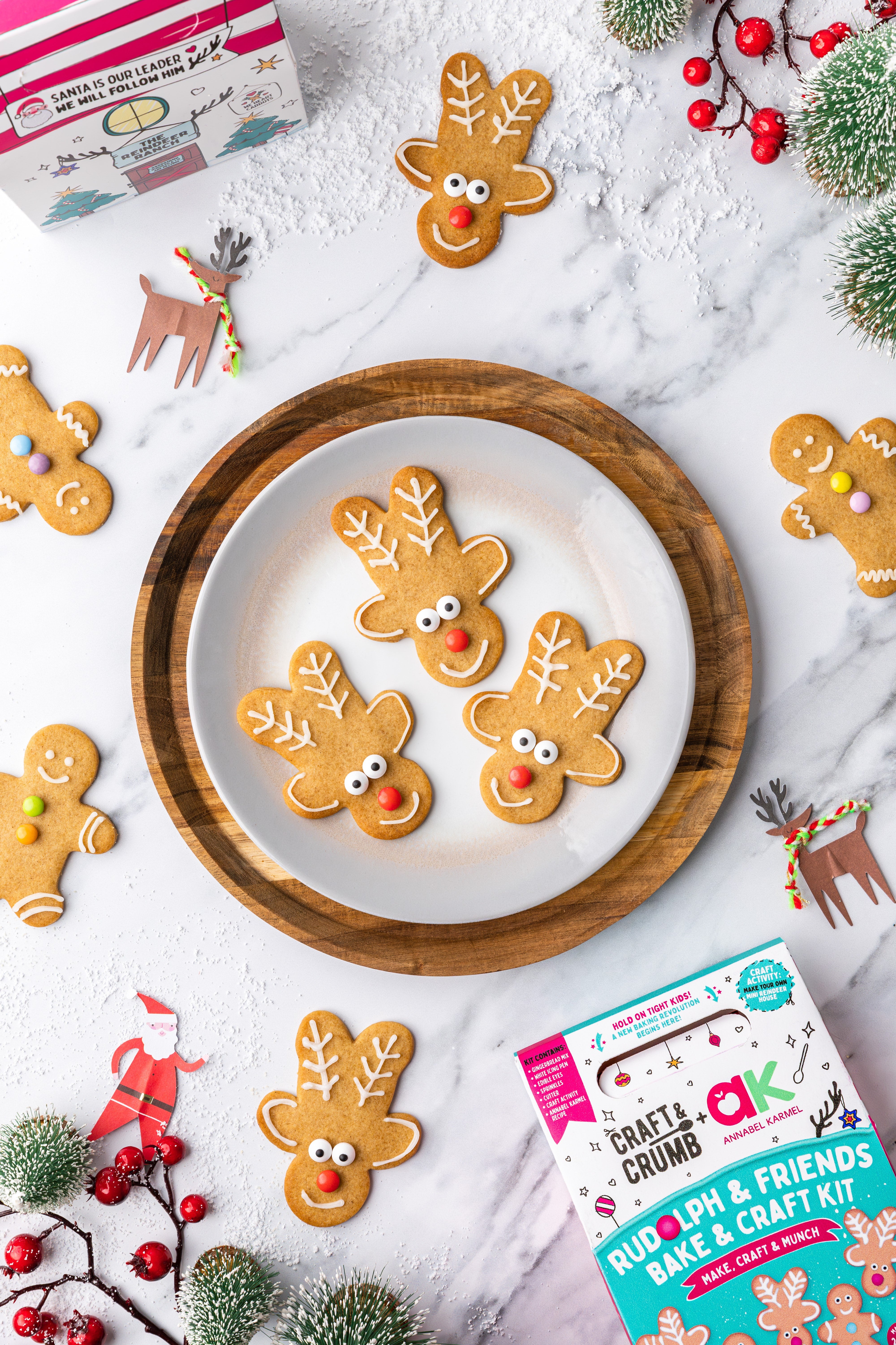 Gingerbread cookies shaped like reindeer on a plate, surrounded by holiday decorations and baking kits.
