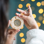 Person holding a decorated cookie ornament with a red and white string, against a background of blurred yellow lights.
