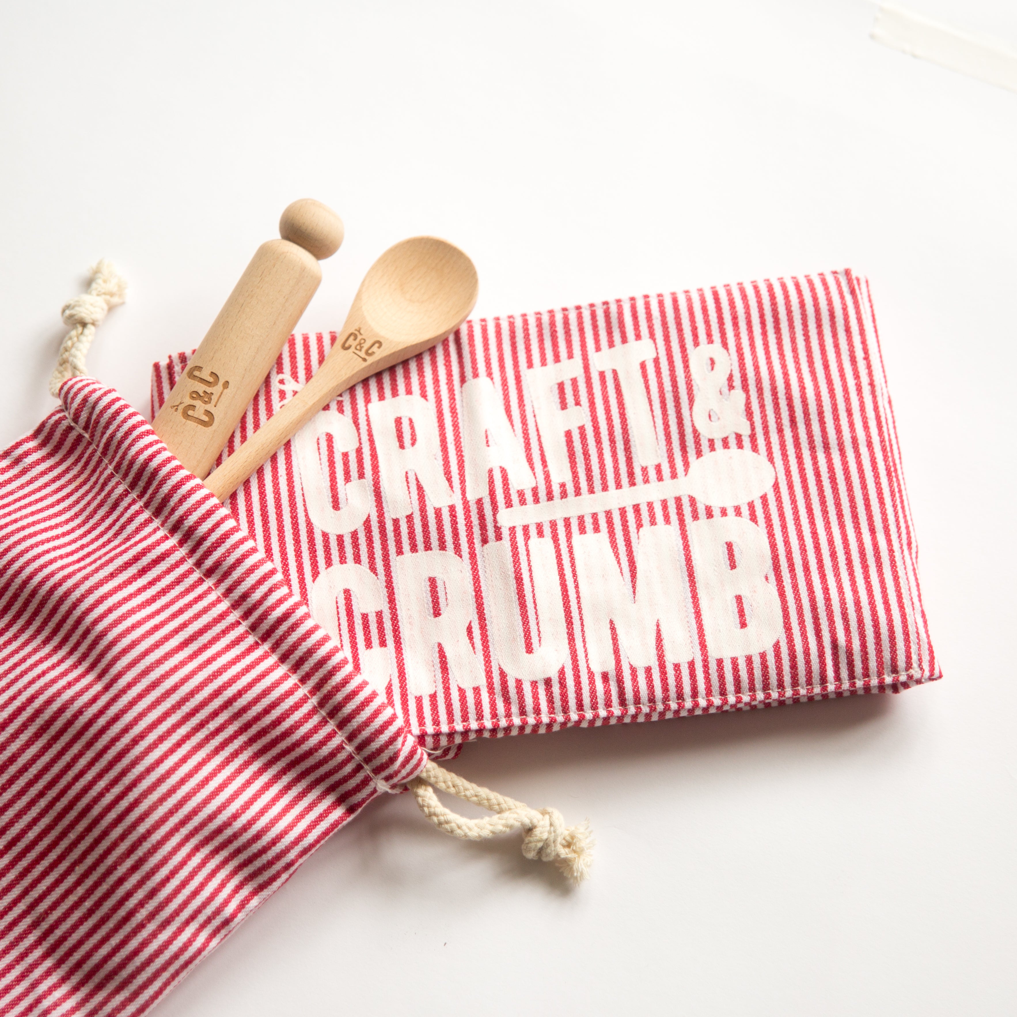 Red striped pouch with wooden spoon and stamper, next to a matching cloth with "Craft & Crumb" text.