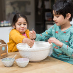 Two children mixing ingredients in a large bowl on a kitchen table, with small bowls and a carton nearby.
