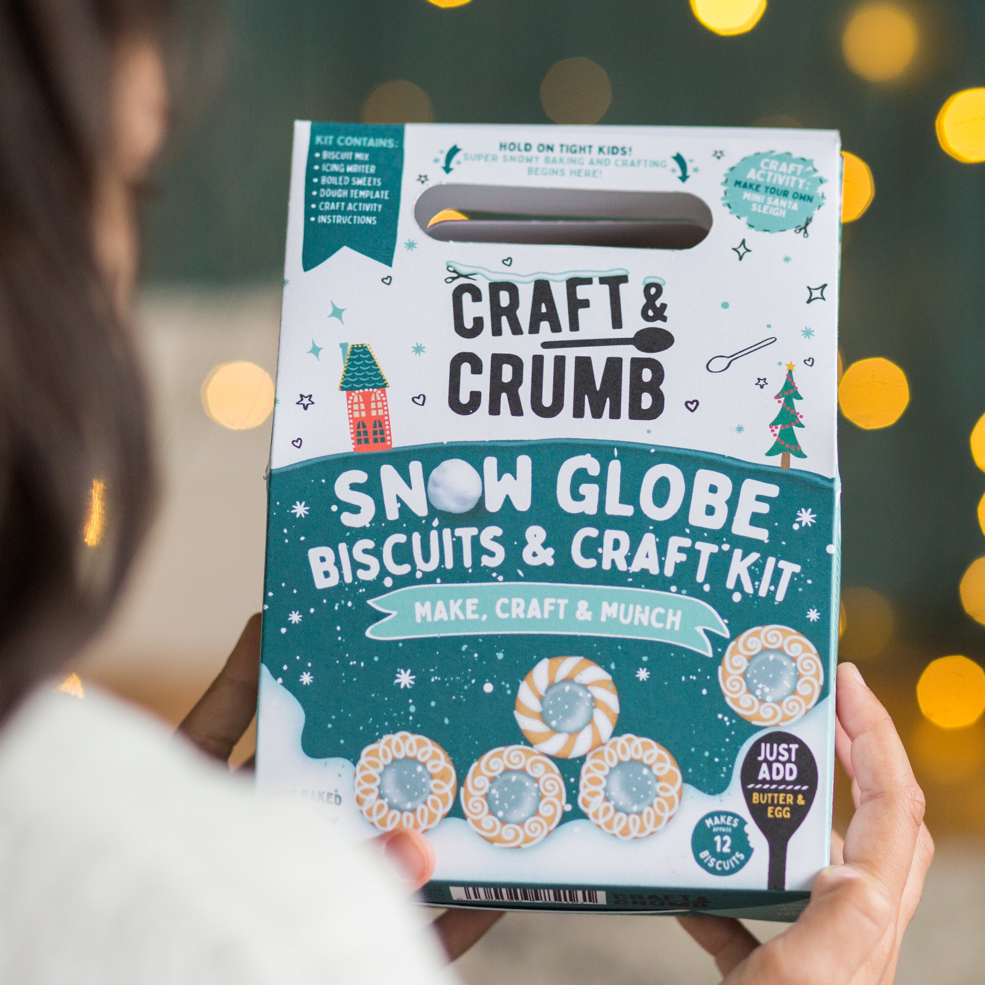 Person holding a "Craft & Crumb" Snow Globe Biscuits & Craft Kit box with festive design and lights in the background.