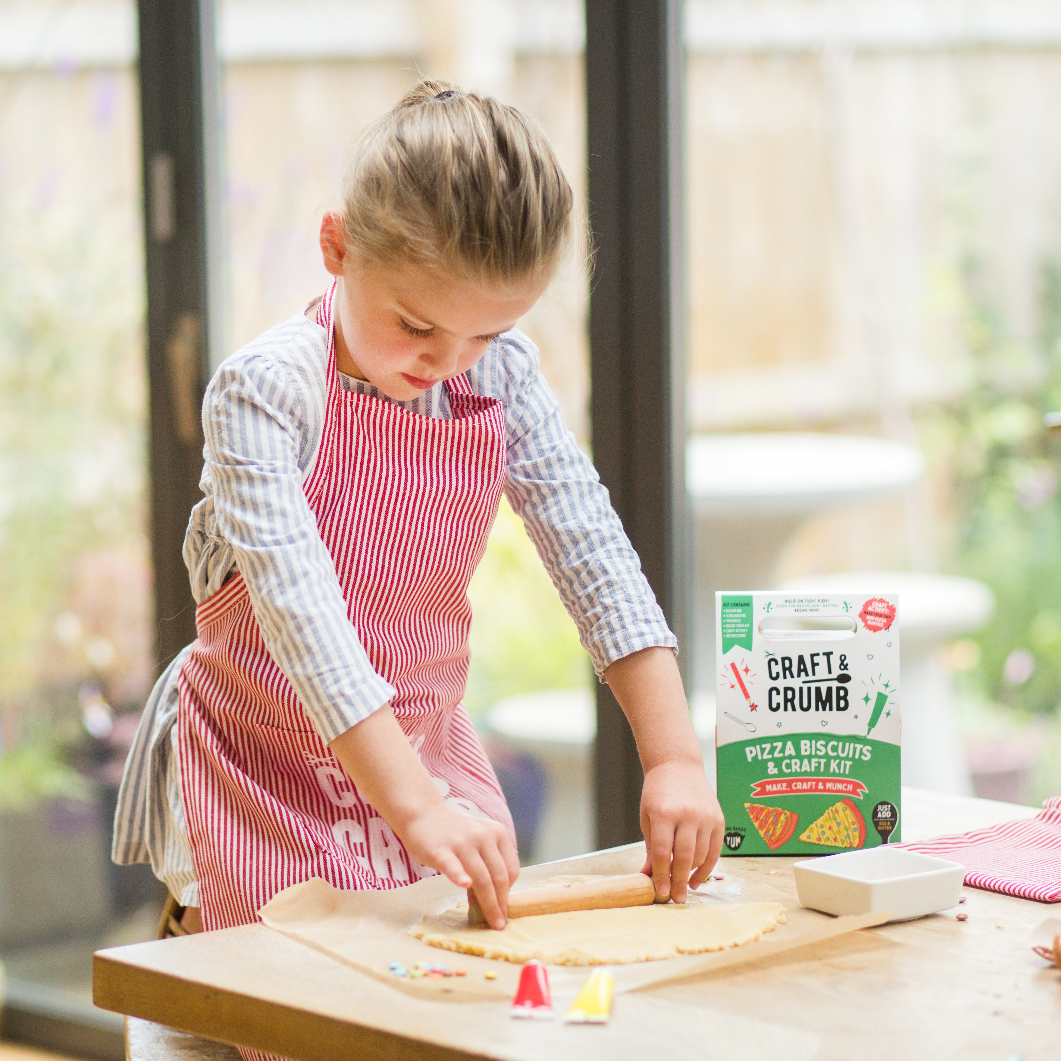 Young girl in a red apron making pizza biscuits with a craft kit on a wooden table.