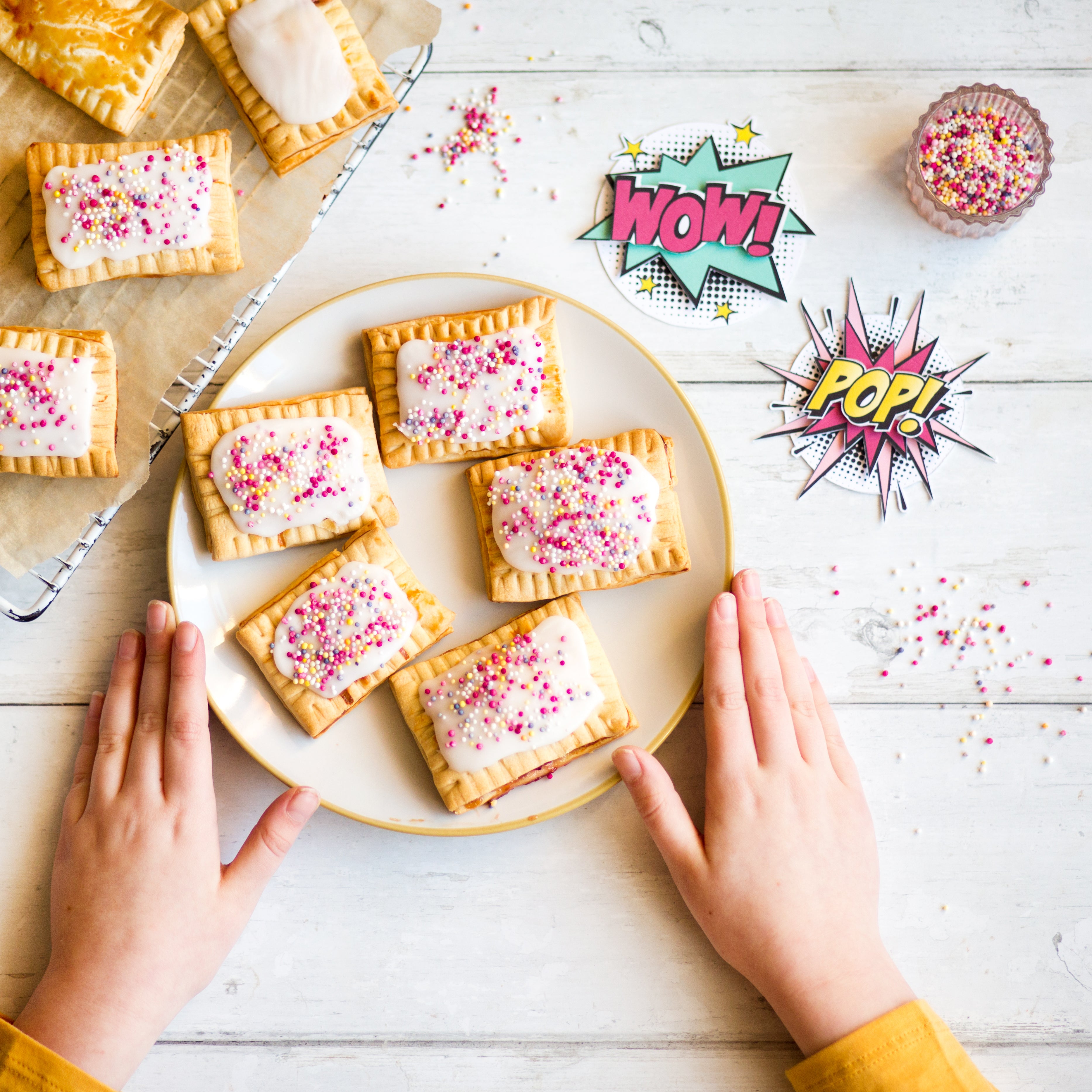 Hands reaching for frosted pastries with sprinkles on a plate, surrounded by comic-style "Wow!" and "Pop!" signs.