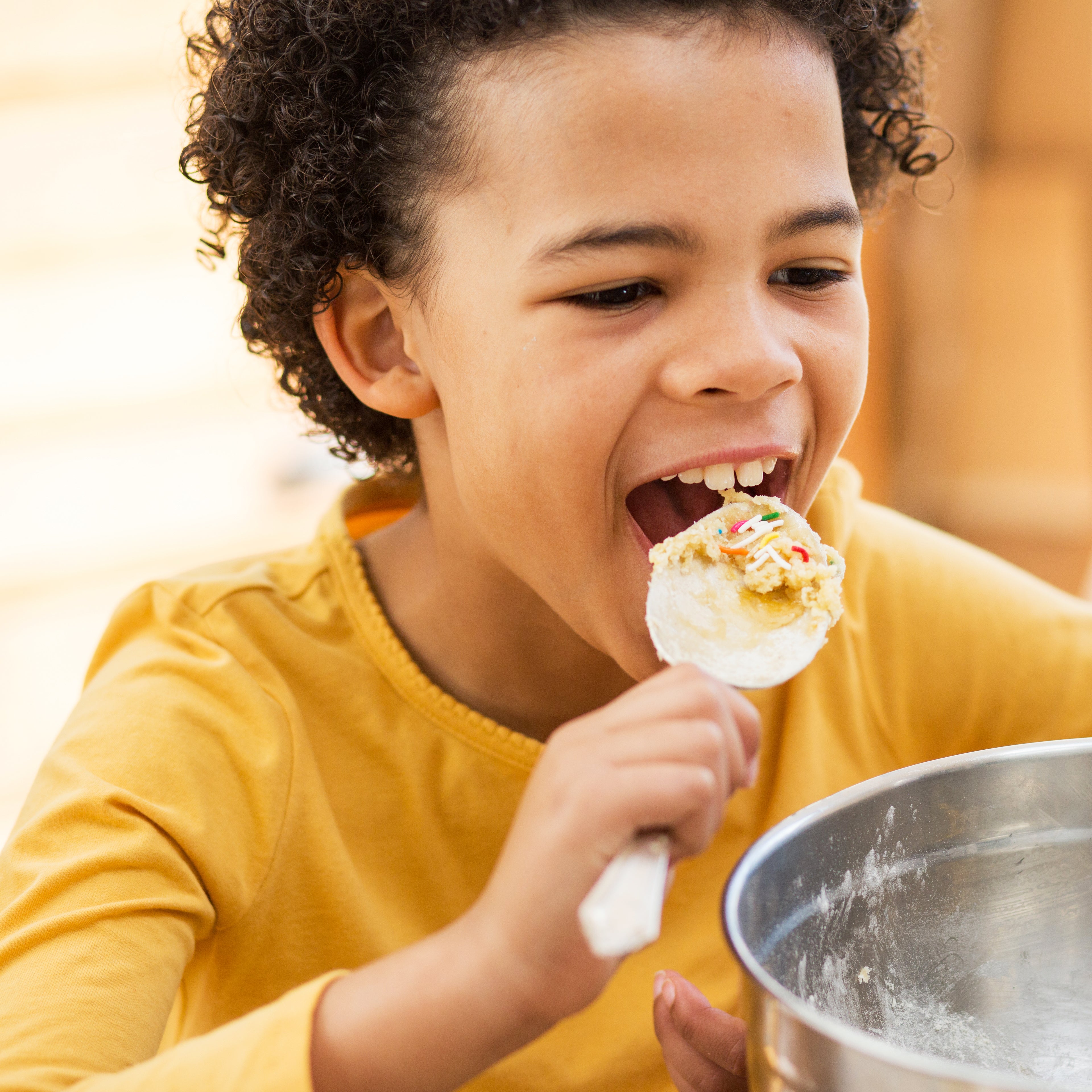 Child enjoying baking with Craft & Crumb kit, licking spoon with cake batter and sprinkles.
