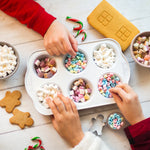Children's hands reaching for colorful candies and marshmallows in a muffin tin, with gingerbread cookies nearby.