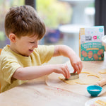 Child cutting cookie dough with a cutter on a table, Easter-themed baking kit in the background.