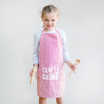 Young girl in a red-striped apron holding a wooden spoon and rolling pin, smiling against a plain background.