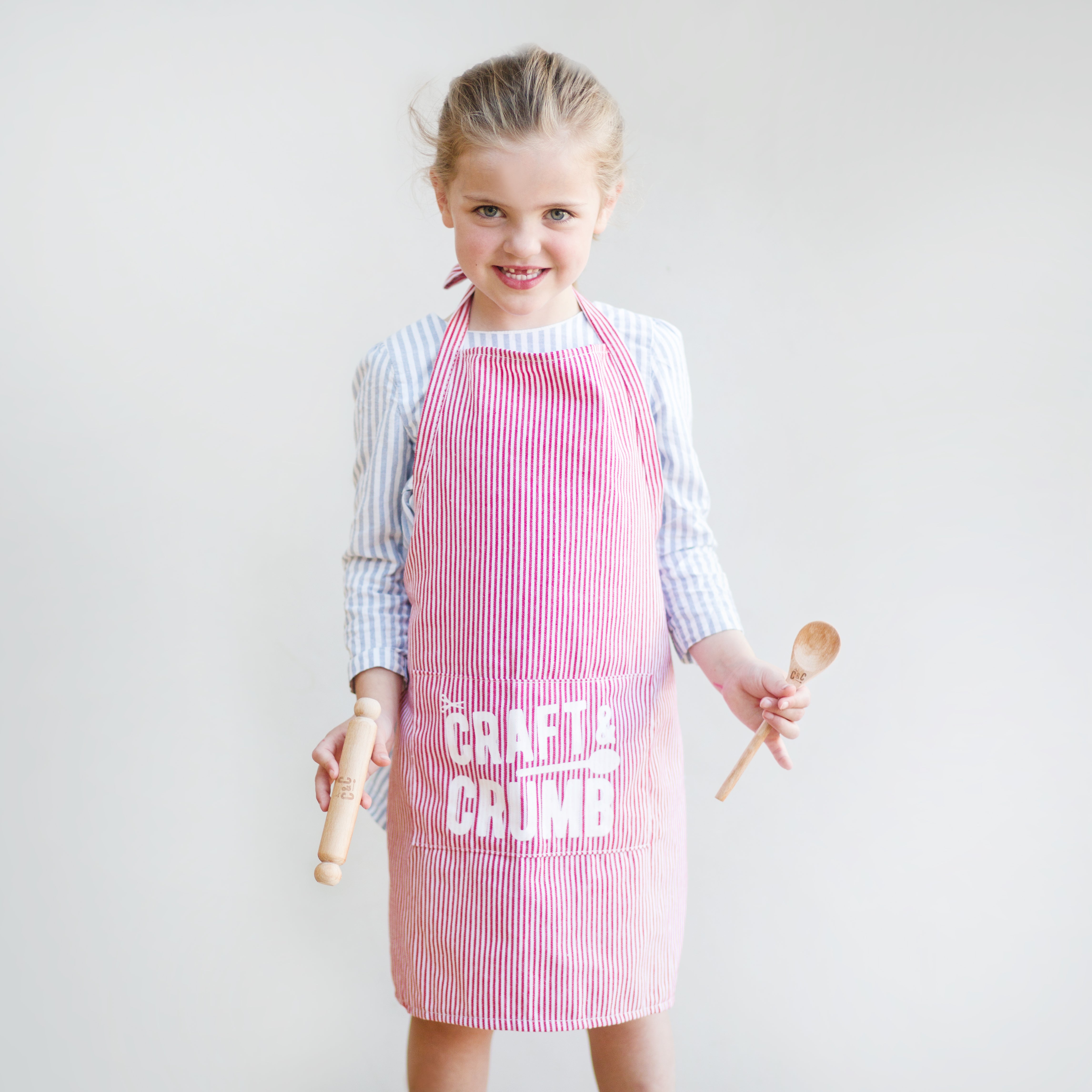 Young girl in a red-striped apron holding a wooden spoon and rolling pin, smiling against a plain background.