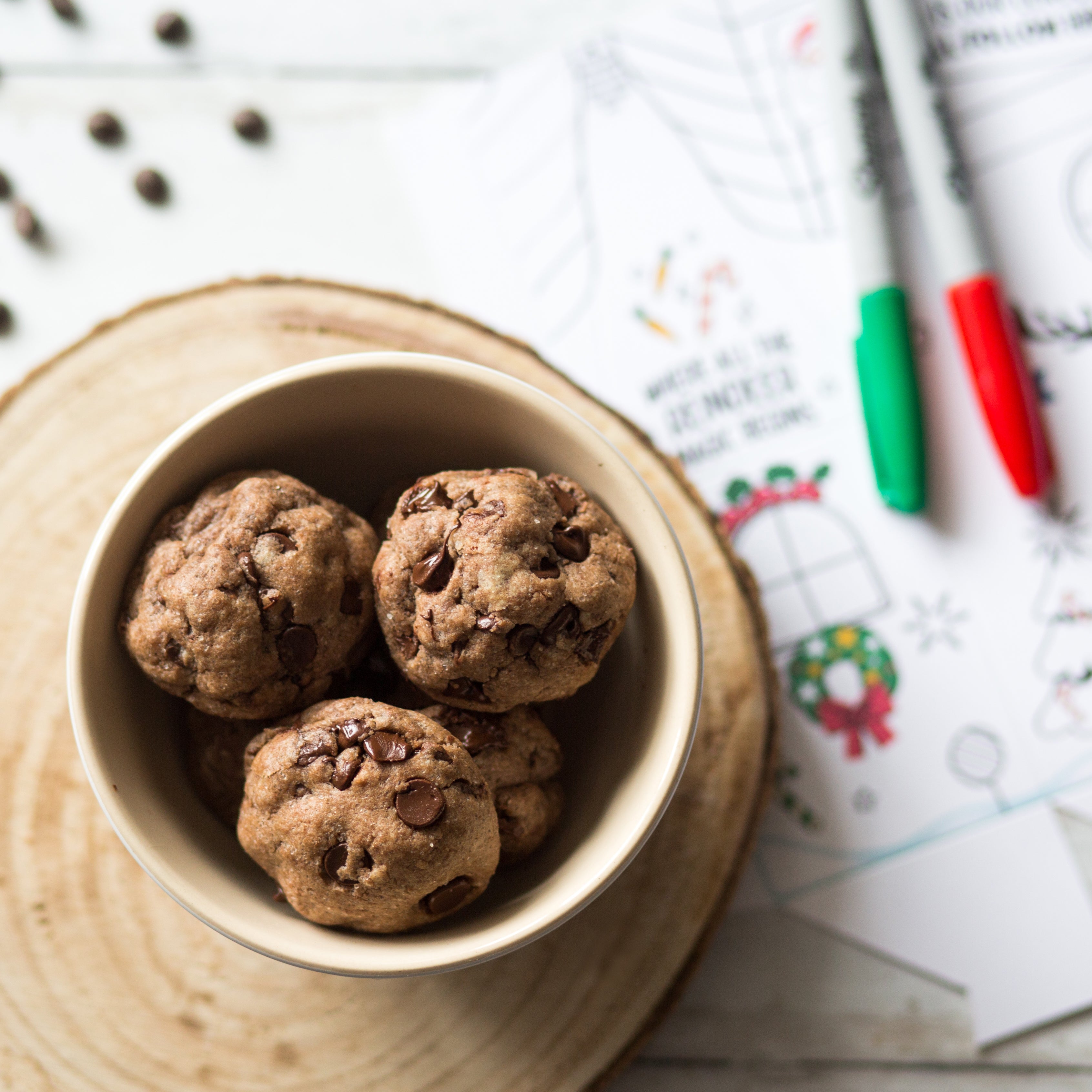 Bowl of chocolate chip cookies on a wooden slab with holiday-themed coloring pages and markers in the background.