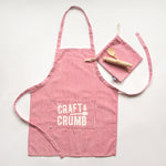 Red and white striped apron with "Craft & Crumb" text, matching pouch, and wooden spoon on a light background.