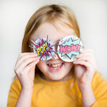 Child smiling, holding comic-style "POP!" and "WOW!" signs over eyes, wearing a yellow shirt.