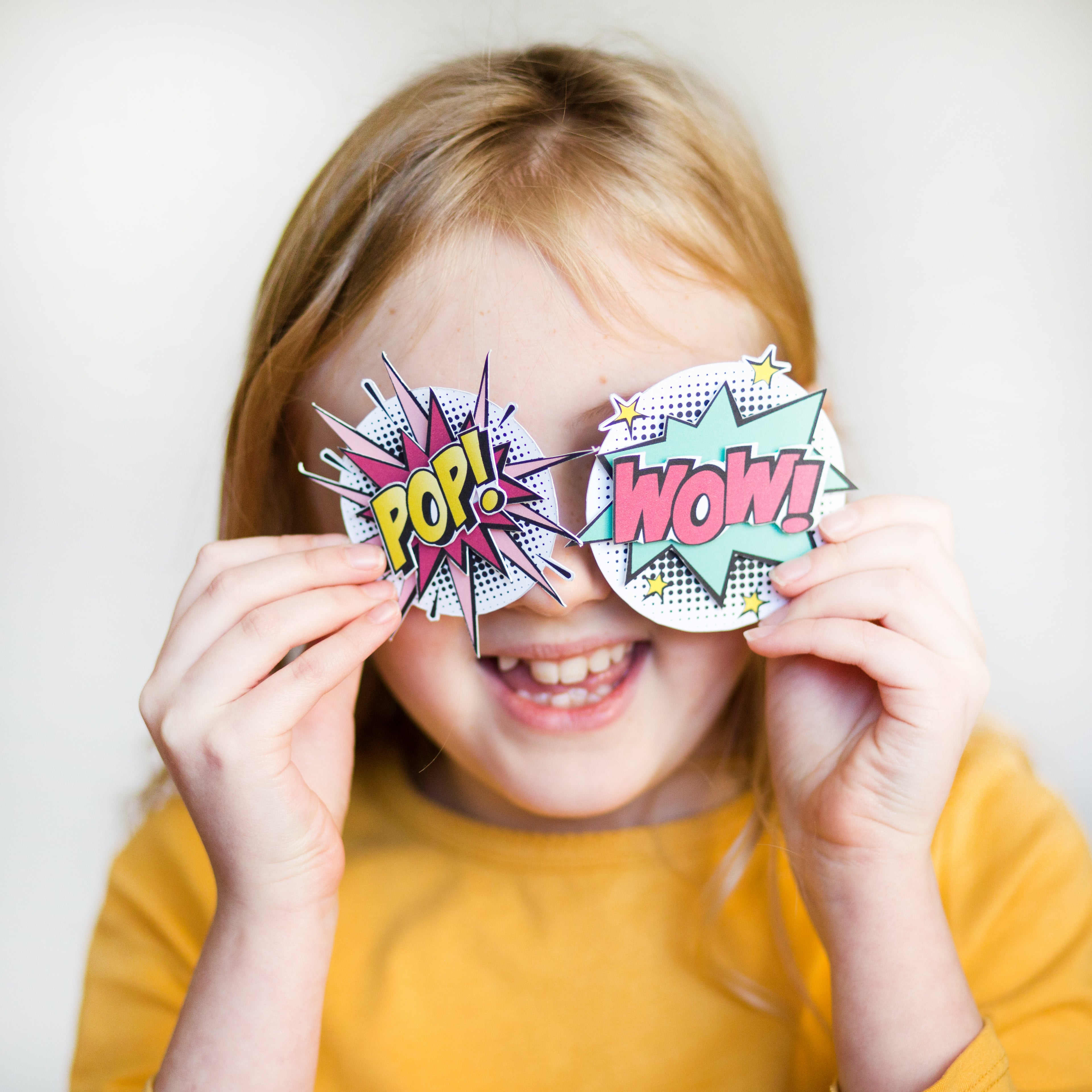 Child smiling, holding comic-style "POP!" and "WOW!" signs over eyes, wearing a yellow shirt.