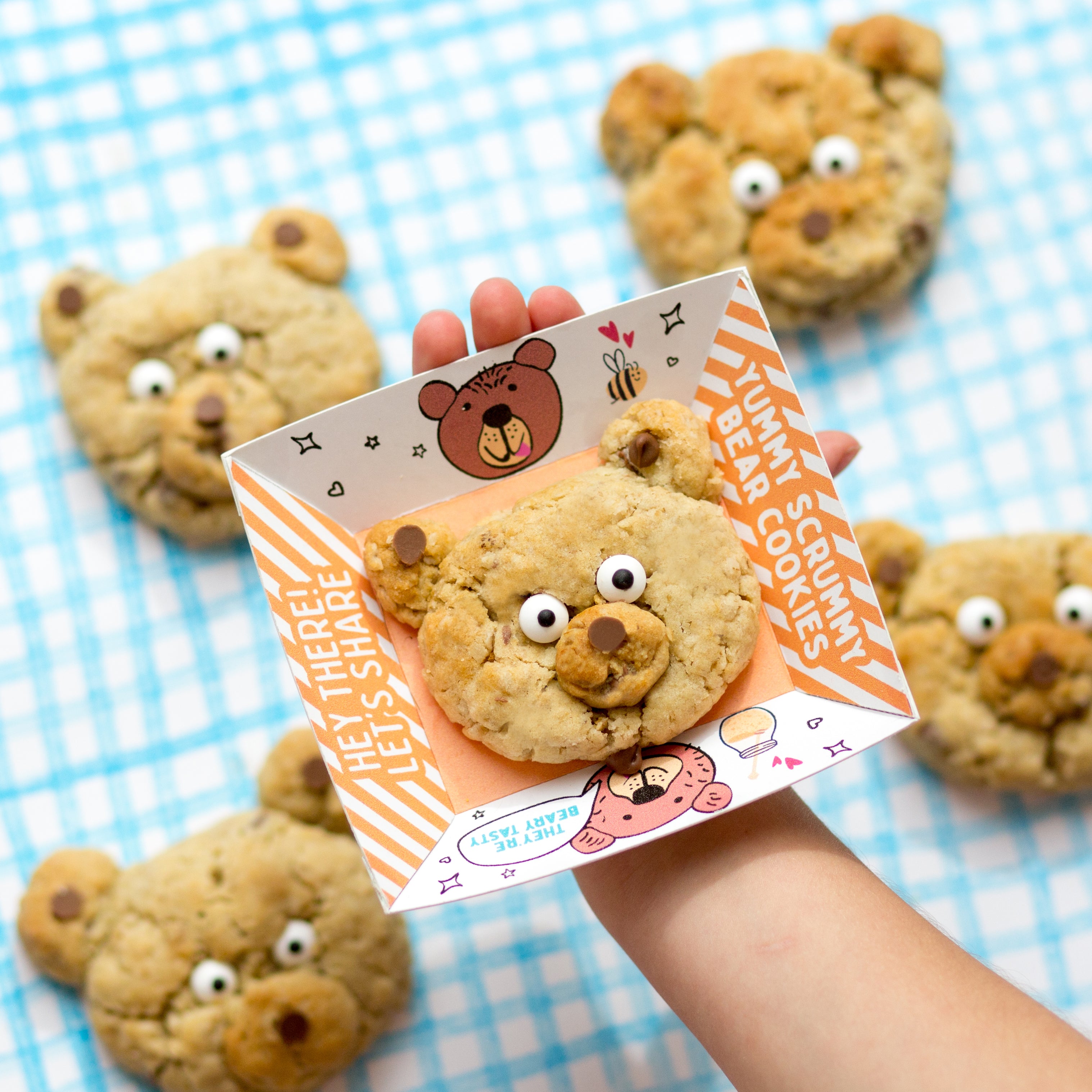 Child holding a bear-shaped cookie on a card, surrounded by more cookies on a blue checkered cloth.