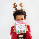 Child with reindeer antlers holding a festive gift box, wearing a red sweater with a reindeer design.