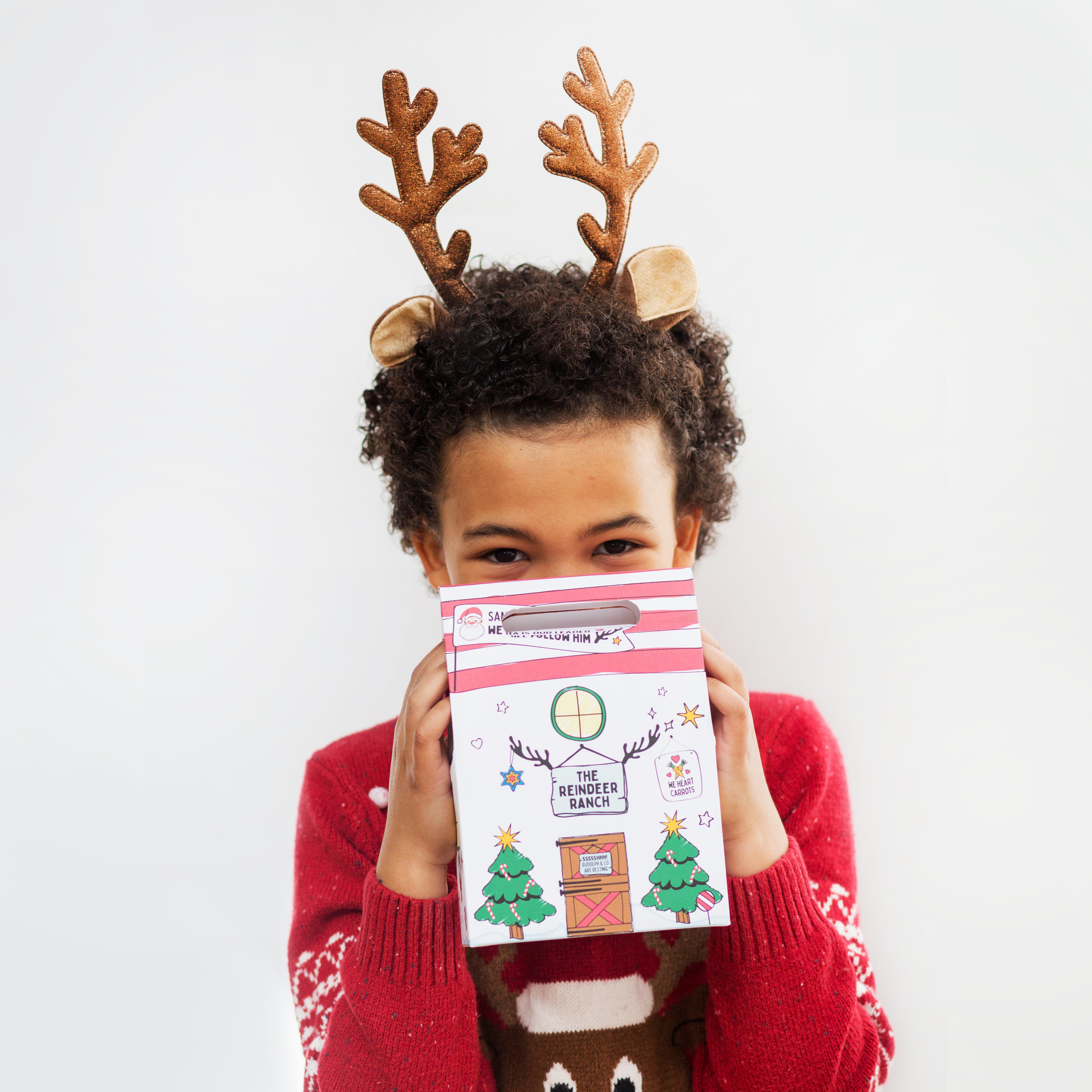Child with reindeer antlers holding a festive gift box, wearing a red sweater with a reindeer design.