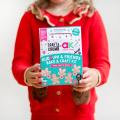 Child holding a "Rudolph & Friends Bake & Craft Kit" in a festive red outfit.