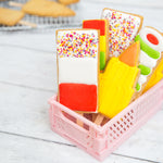 Colorful, decorated cookies shaped like popsicles and pencils in a pink basket on a white wooden surface.