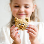Child holding a bear-shaped cookie with chocolate chip eyes and a nose, smiling in the background.
