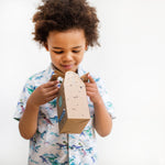 Child in dinosaur shirt holding a cardboard box with a curious expression against a plain background.