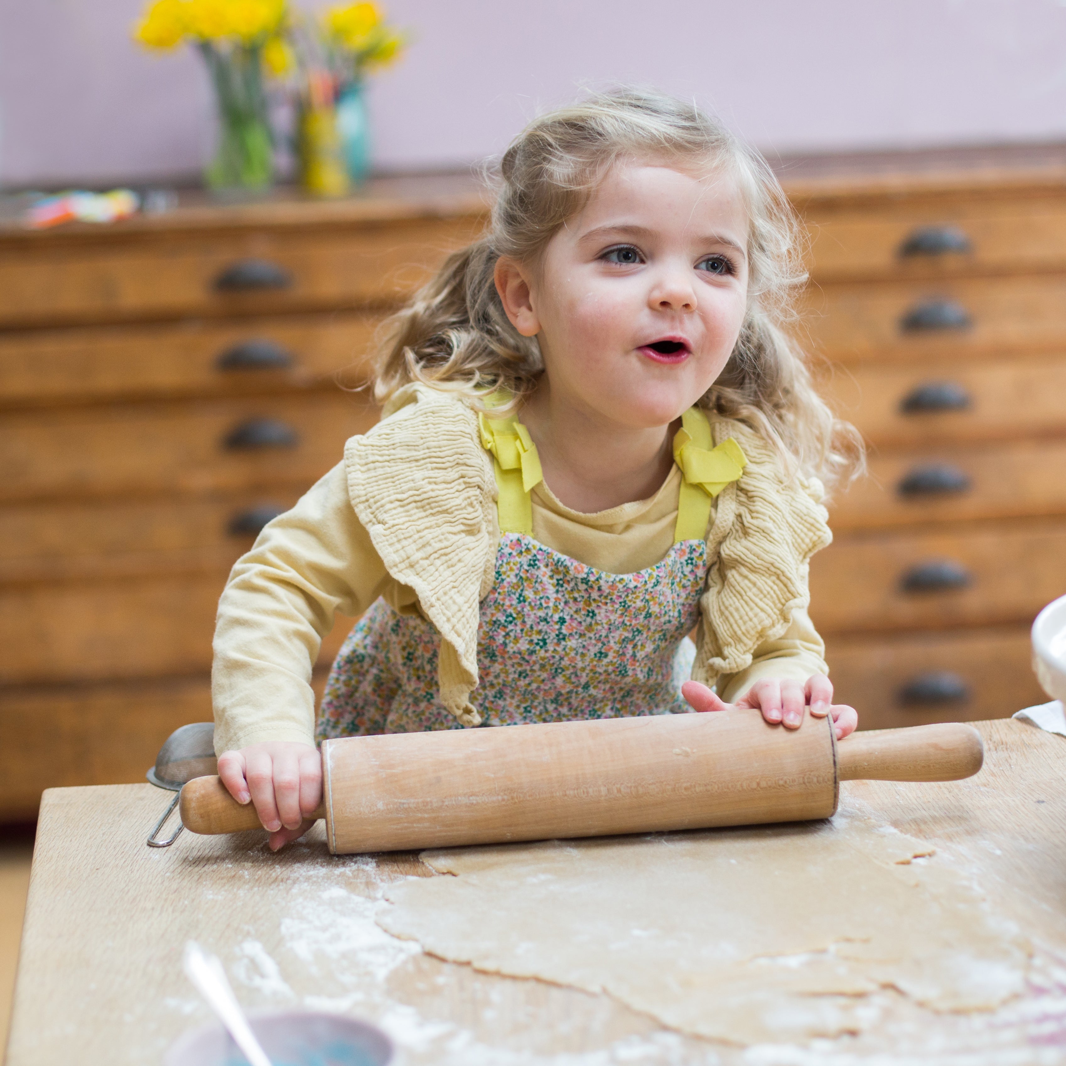 Young girl in a floral apron rolling dough with a pin on a wooden table, with flowers in the background.