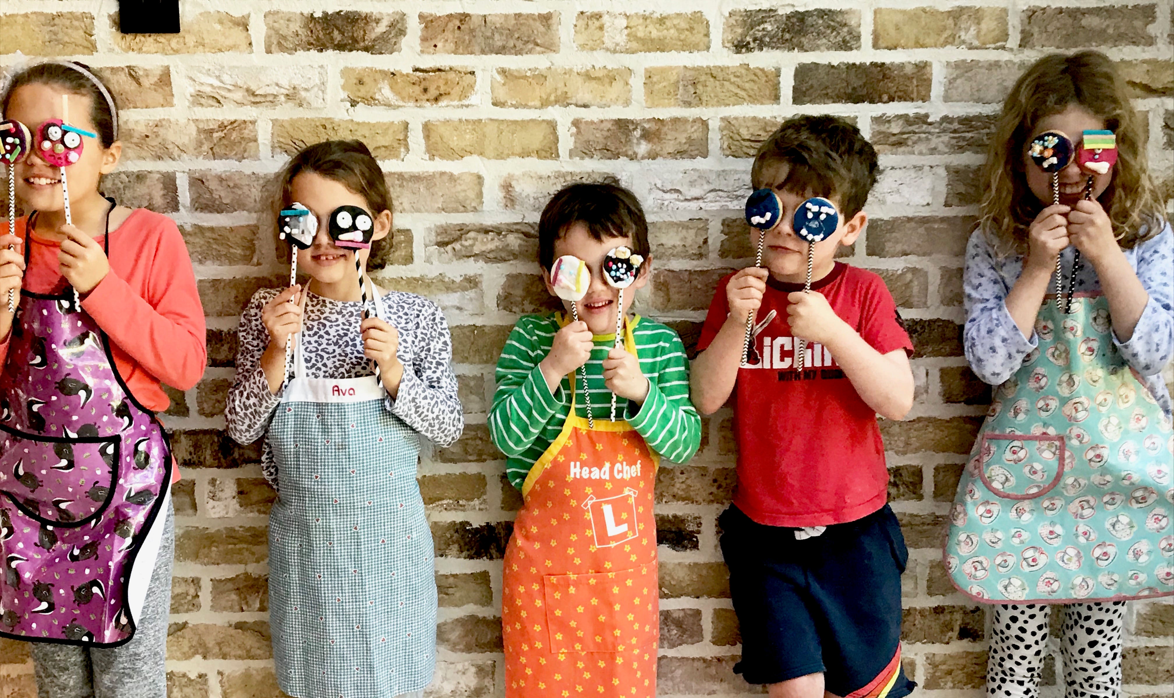 Children wearing colorful aprons holding decorated cake pops in front of their faces, standing against a brick wall, showcasing Craft & Crumb baking kits for kids.
