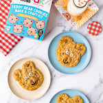 Teddy bear cookies on plates with a baking kit box and milk on a marble surface.