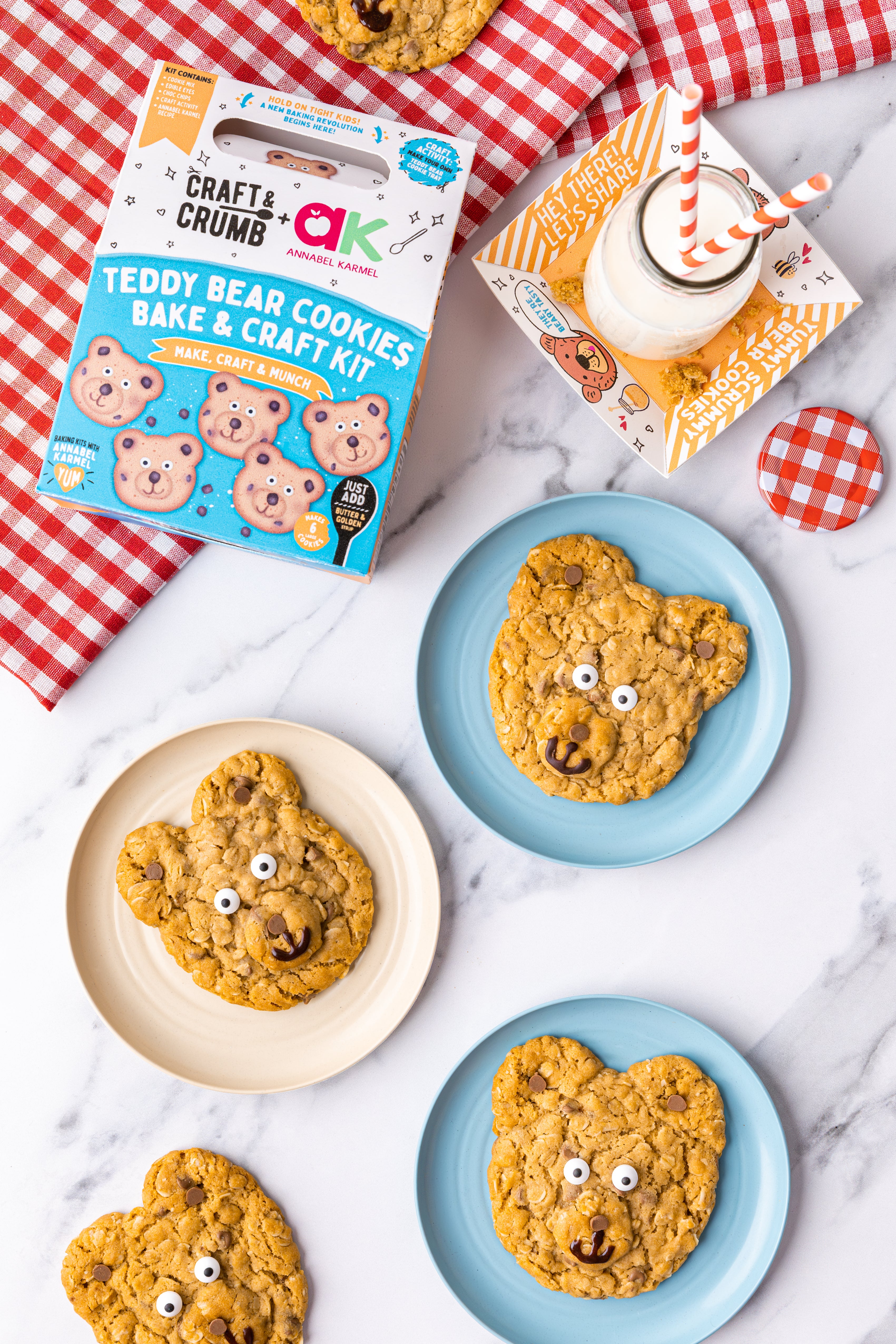 Teddy bear cookies on plates with a baking kit box and milk on a marble surface.