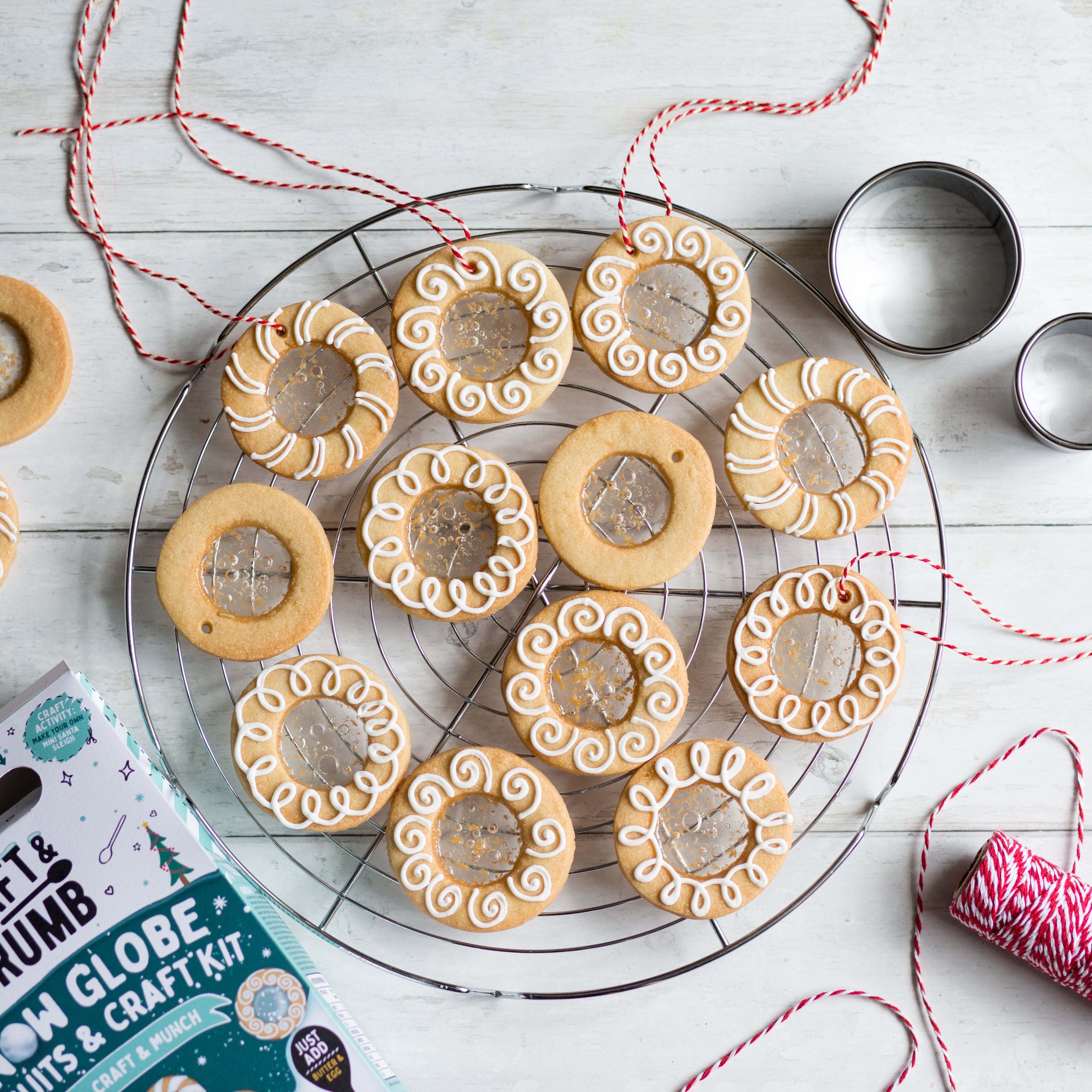 Decorative cookies with icing on a cooling rack, surrounded by baking tools and red twine on a wooden surface.