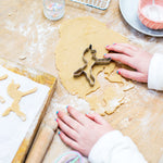 Child's hands using unicorn cookie cutter on dough, with baking tools and cut-out cookies on a wooden surface.