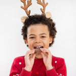 Child in reindeer antlers and red sweater eating a cookie, smiling against a white background.