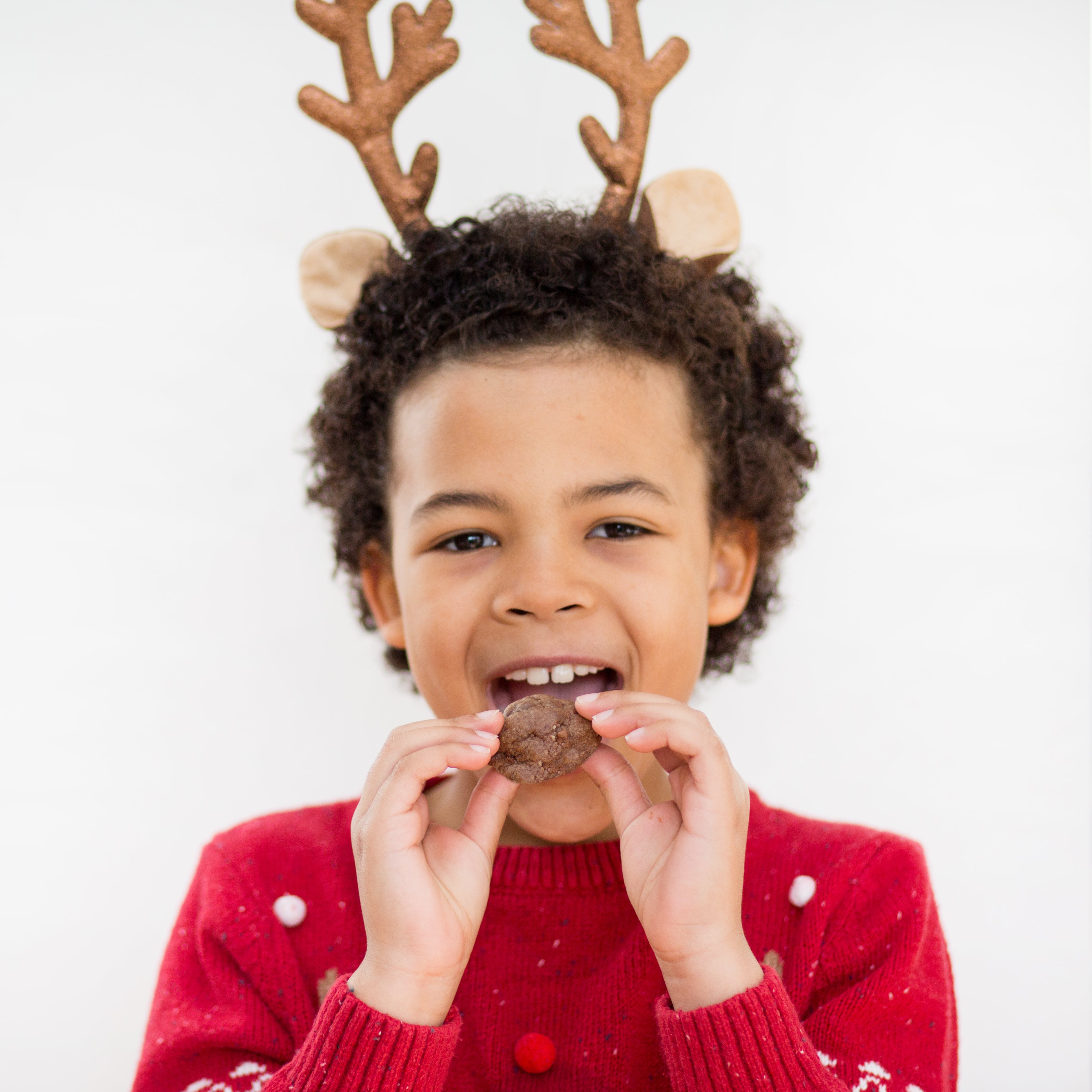 Child in reindeer antlers and red sweater eating a cookie, smiling against a white background.