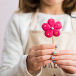 Child holding a pink flower-shaped cookie on a stick, wearing a beige apron and white shirt.