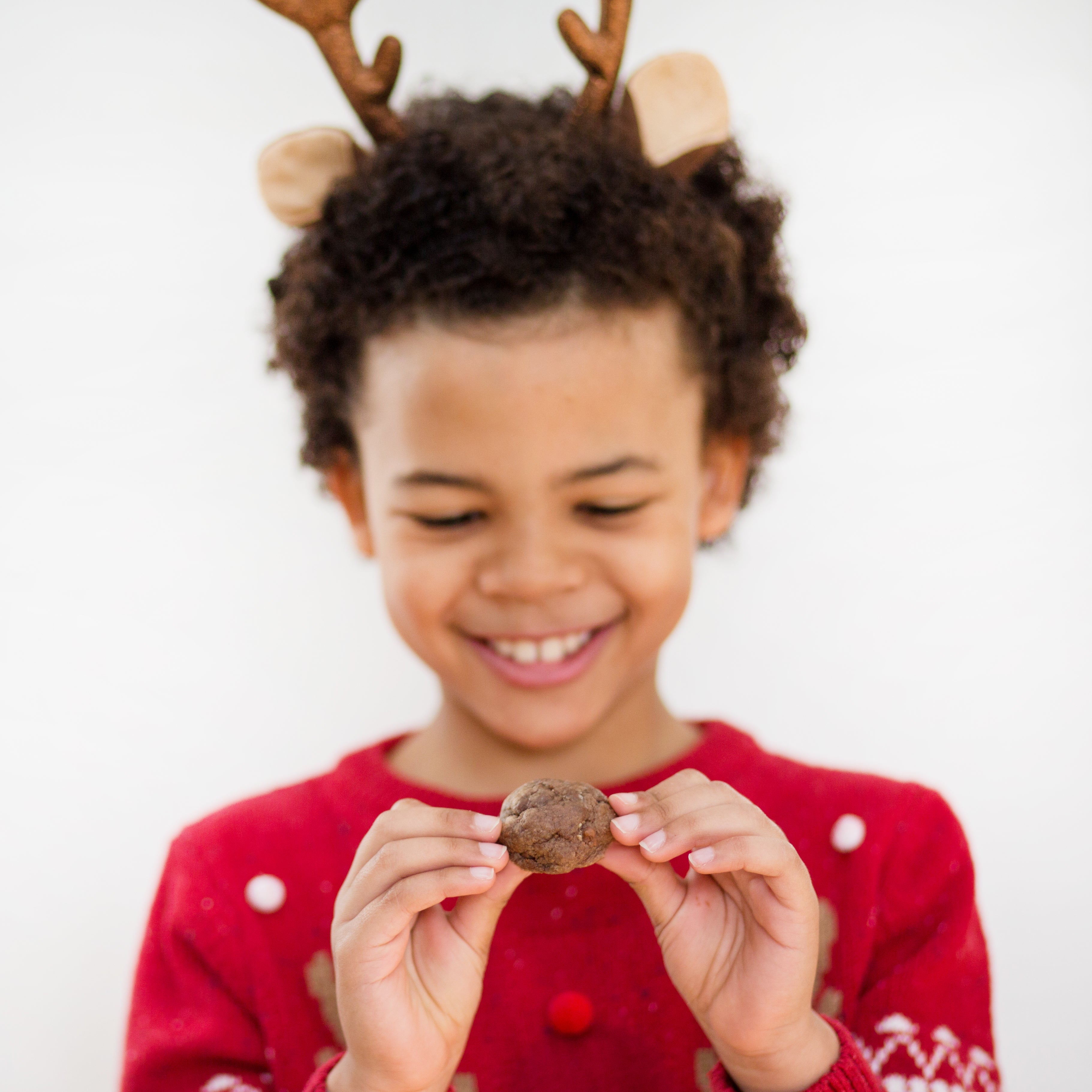 Child in reindeer antlers and red sweater holding a chocolate cookie, smiling against a white background.
