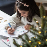 Young girl drawing at a table near a decorated Christmas tree, wearing a white sweater and a headband.