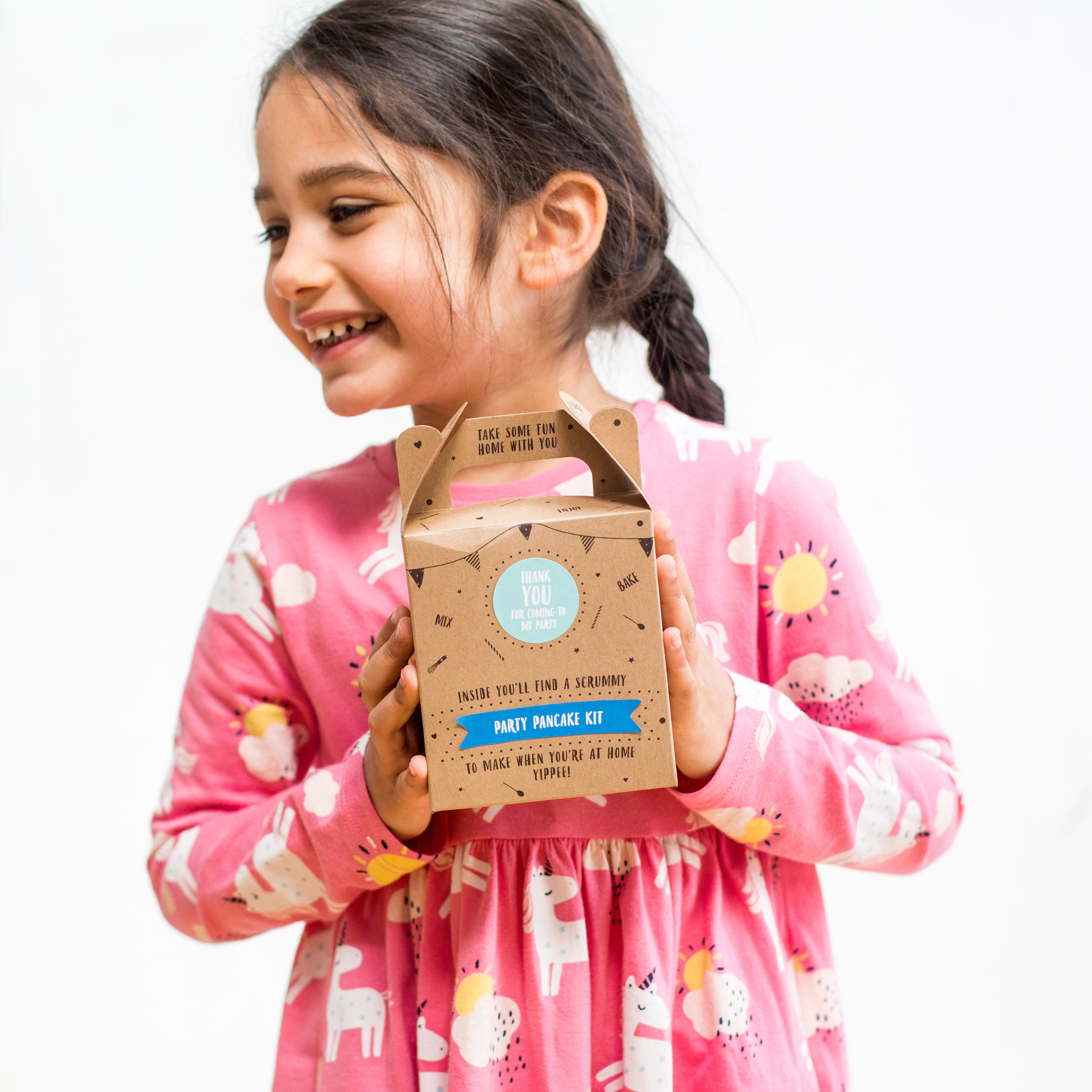 Smiling girl in pink dress holding a party pancake kit box against a white background.